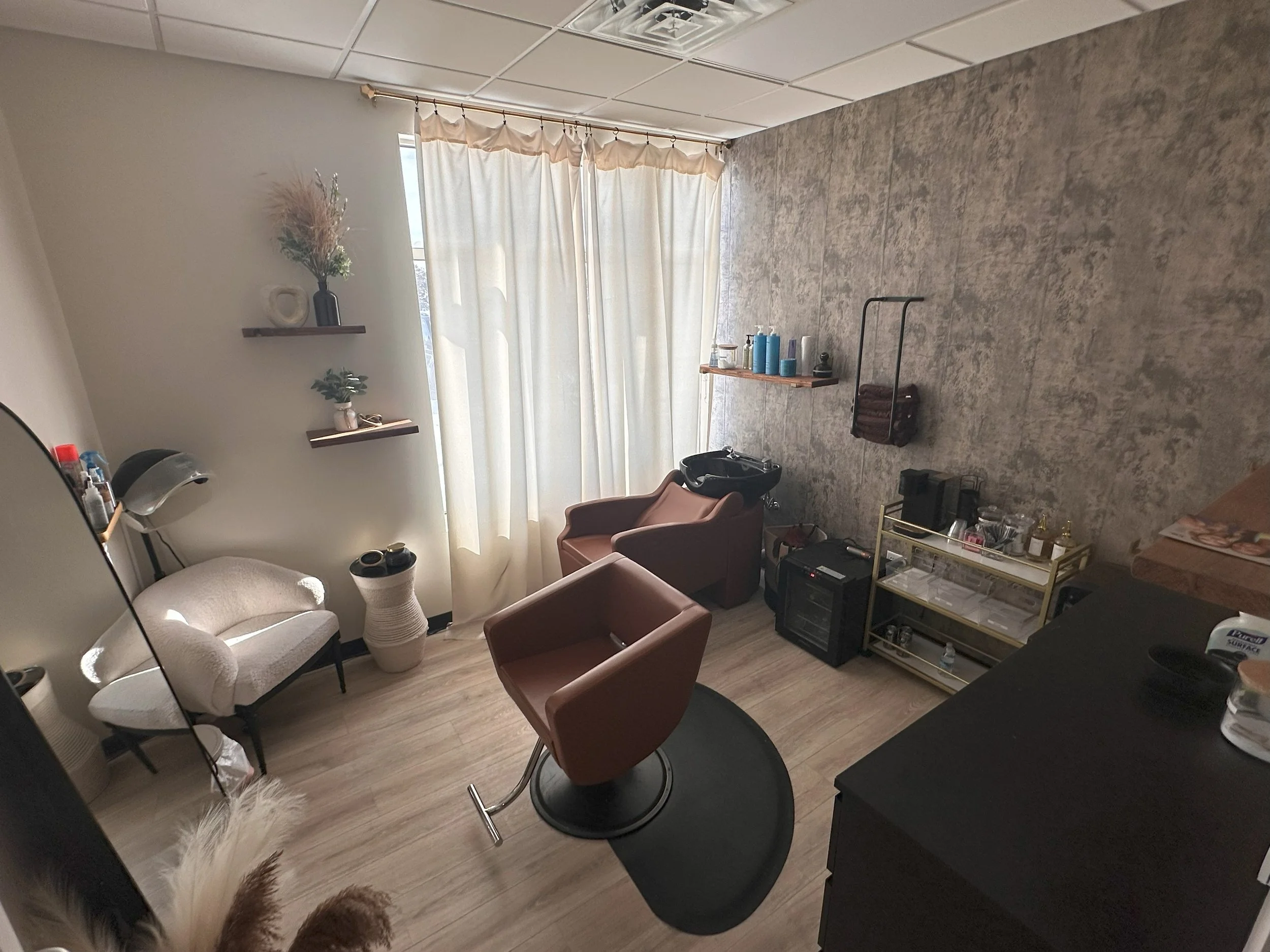 A hair salon room with a hair washing station, styling chair, small table, wall shelves with decorative items, a window with cream curtains, and a variety of salon products and equipment.
