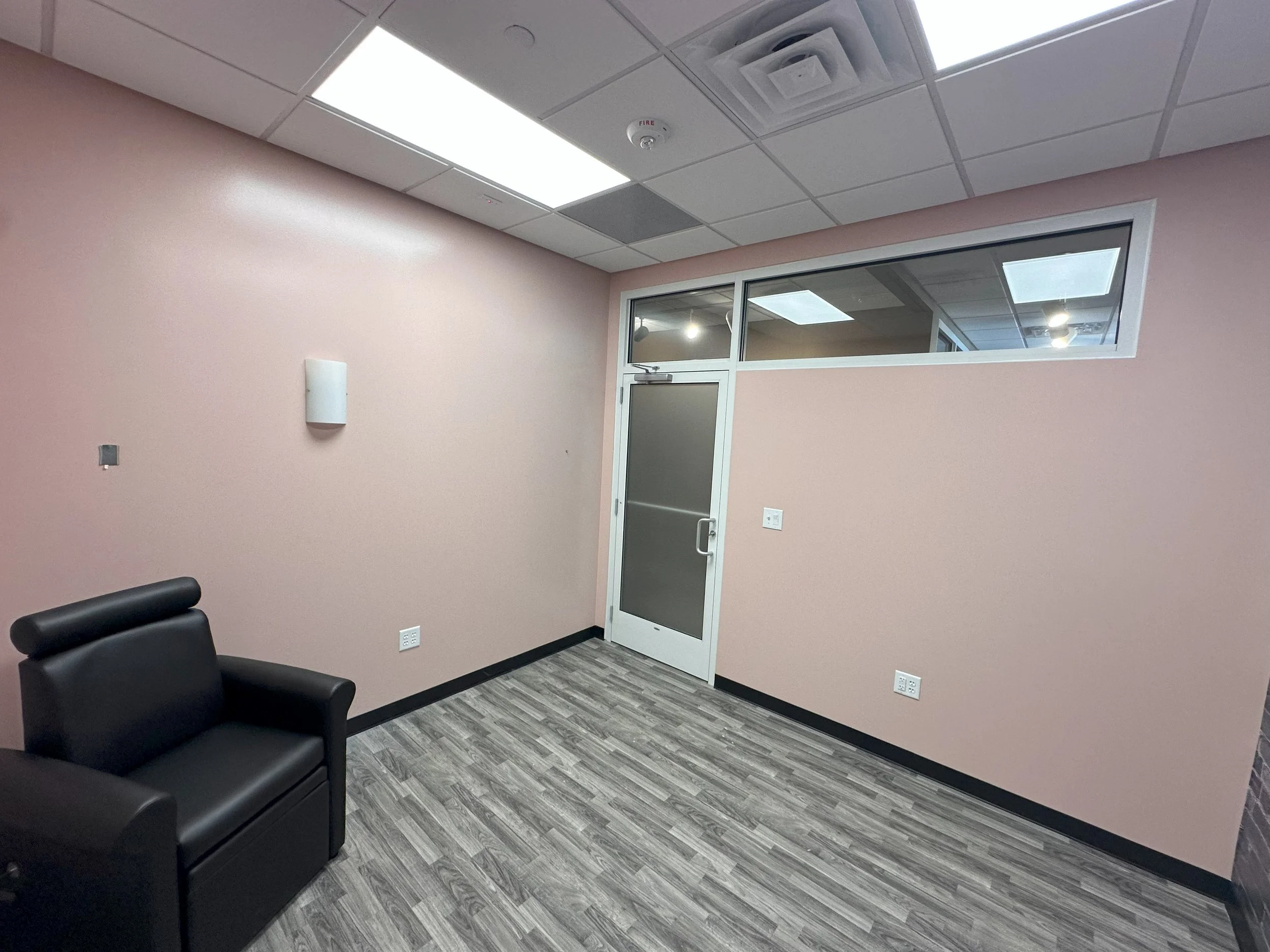 Empty office room with a black chair, pink walls, a glass door, and a high horizontal window. Ceiling has panel lights and ventilation system.