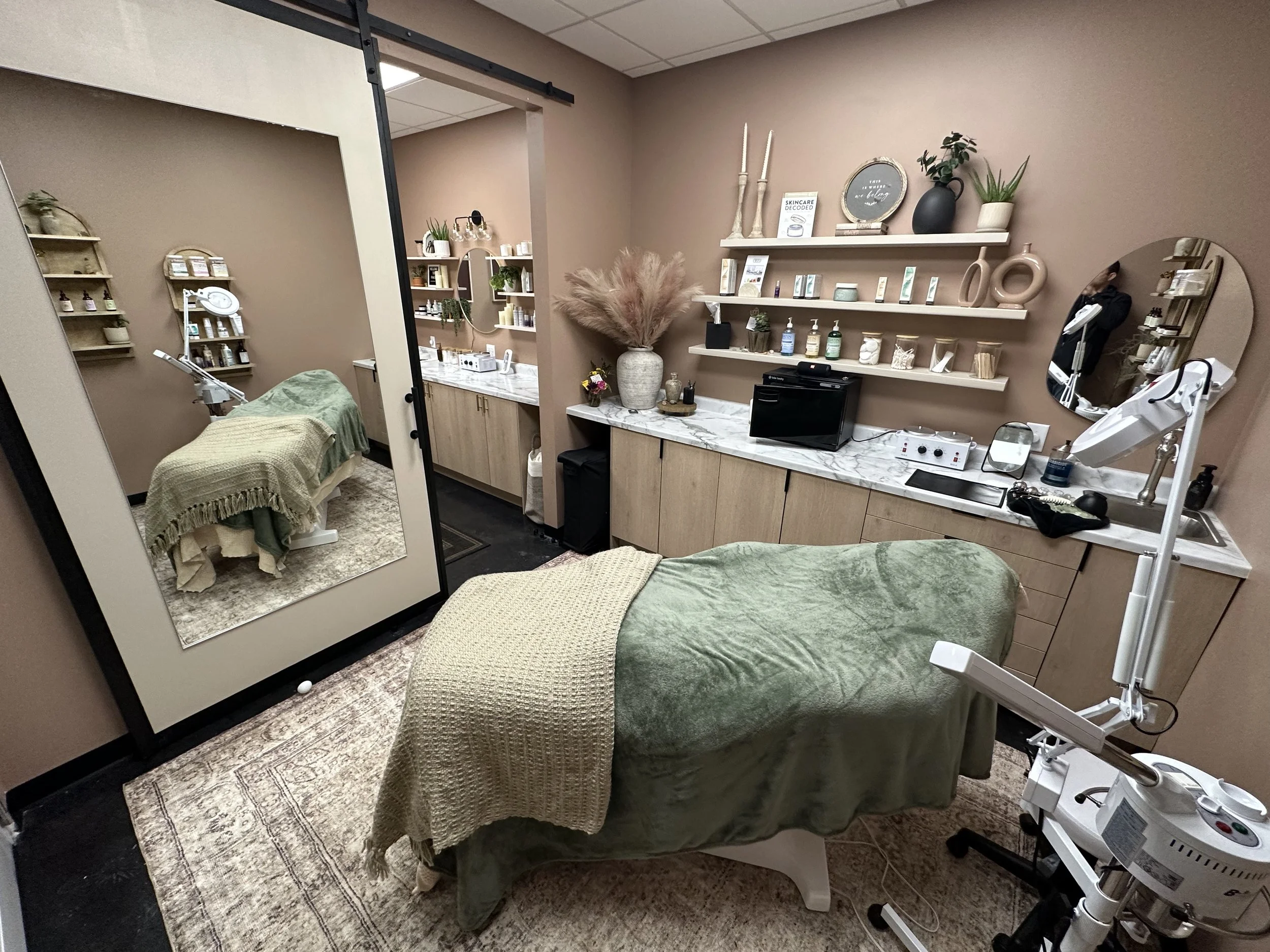 A spa or treatment room with a massage bed covered in a green towel, a large mirror, shelves with skincare products, and various treatment equipment and decor.
