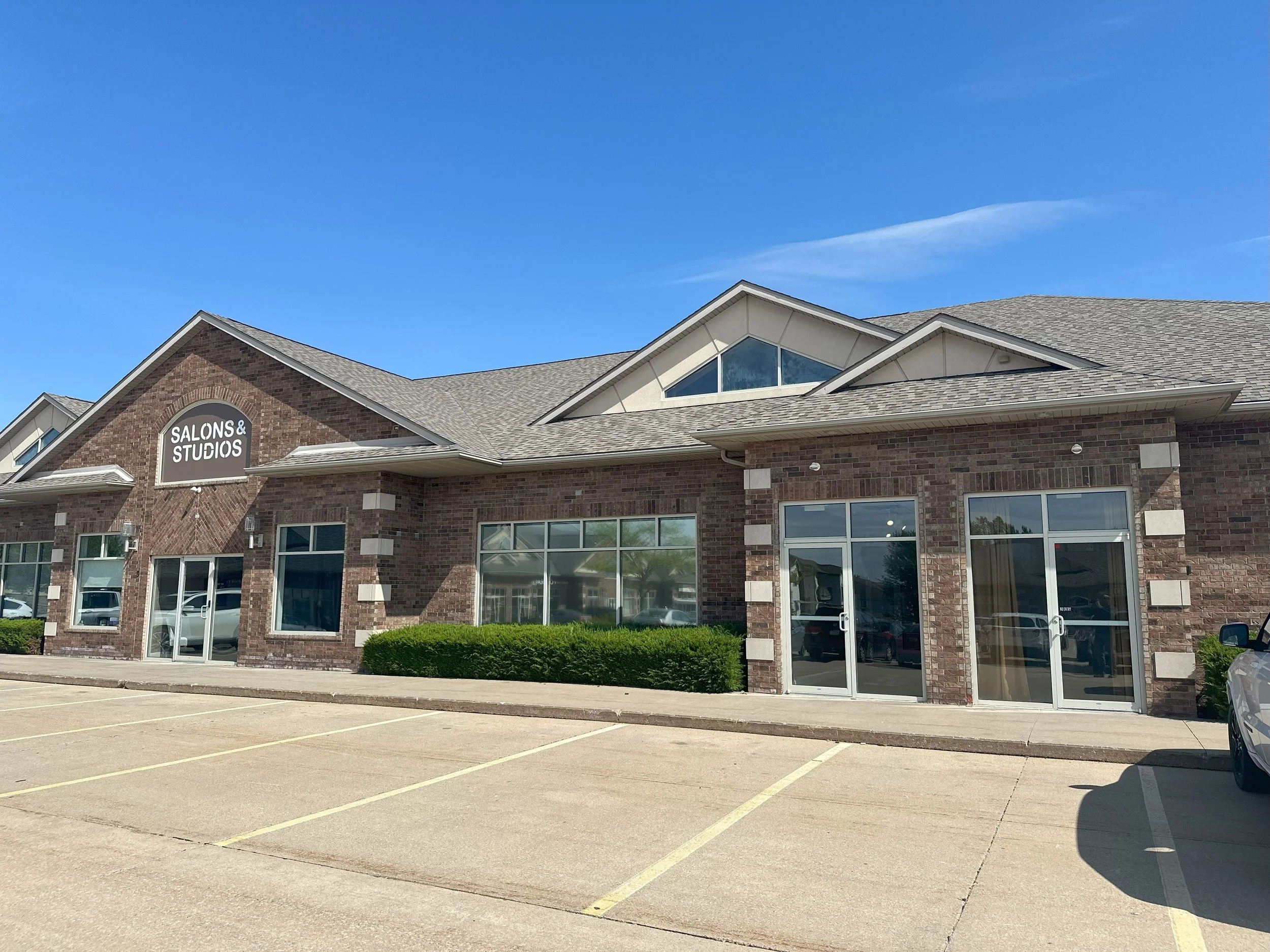 A brick building with a sign that reads 'Salons & Studios' under a gable roof, with large windows and glass doors, and an empty parking lot in front under a blue sky.