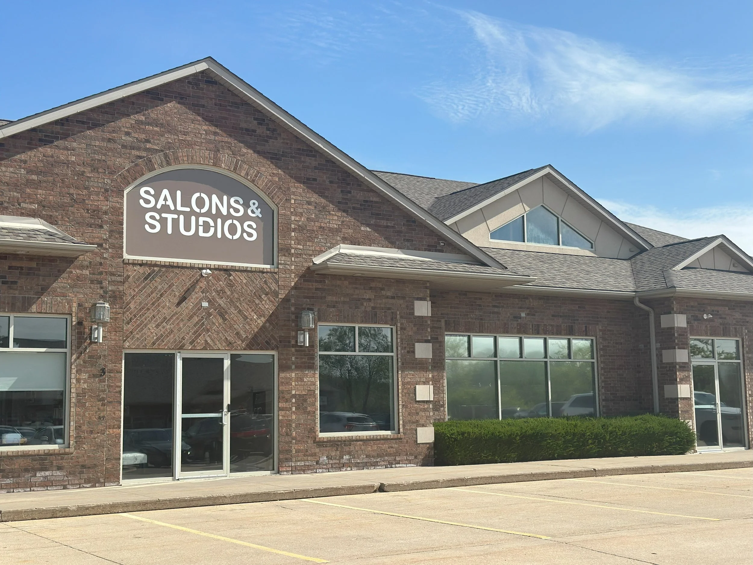 Exterior of a brick building with a sign that reads 'Salons & Studios', large windows, a concrete sidewalk, and an empty parking lot under a blue sky with scattered clouds.