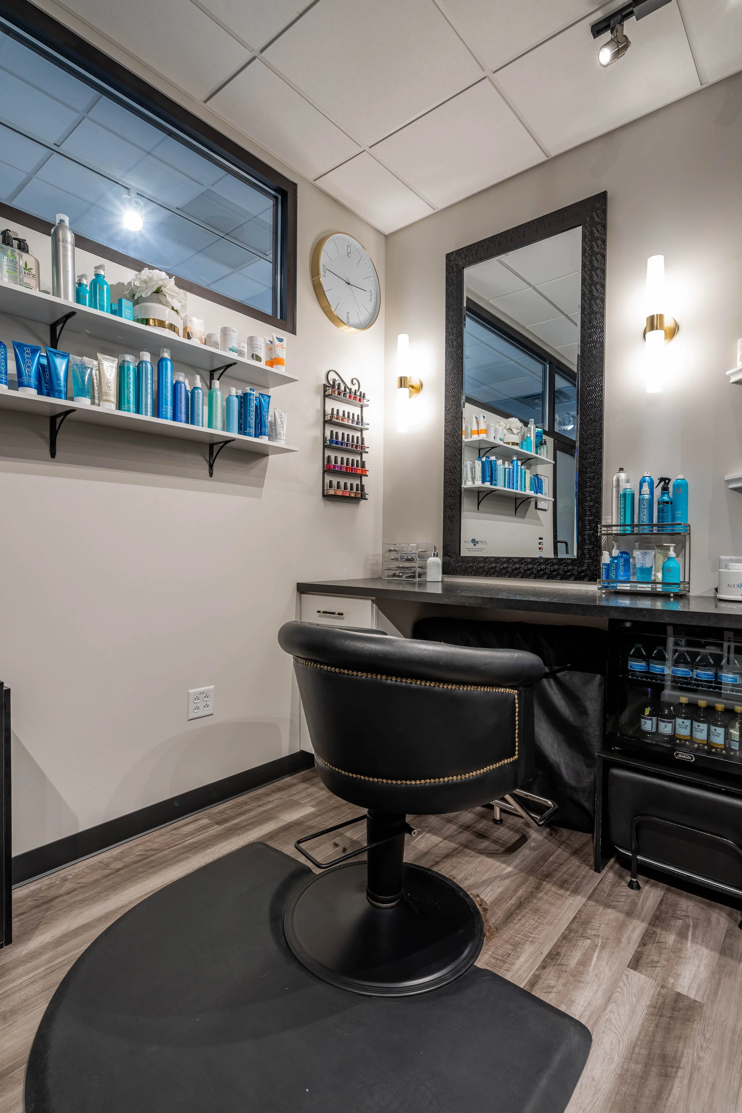Salon station with black barber chair, large mirror, shelves with hair products, nail polish display, and a window.