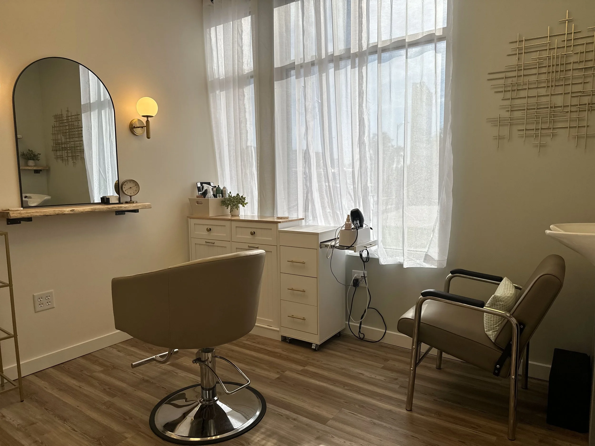 Interior of a hair or beauty salon with a styling chair, a small white cabinet, a mirror on the wall, and a large window with sheer curtains.
