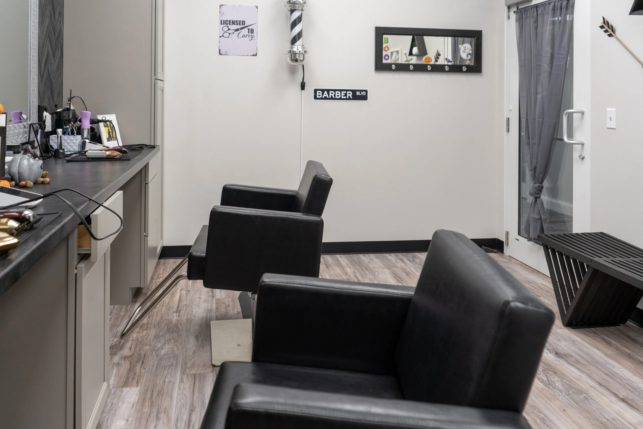 Interior of a hair salon with black chairs, a mirror on the wall, a door with a gray curtain, and various salon tools on the counter.