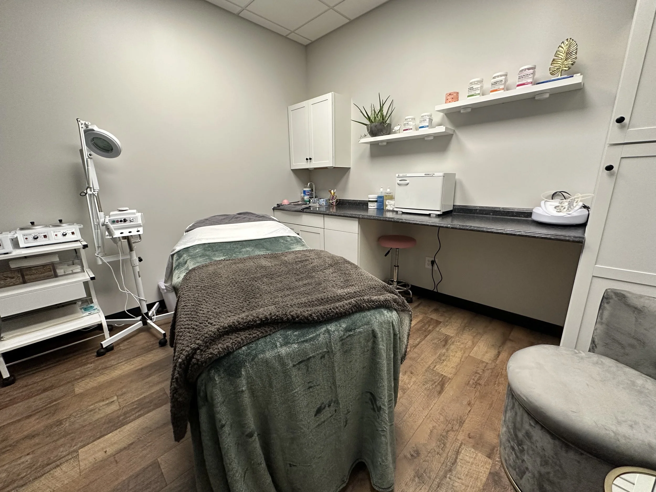 Medical treatment room with a massage or examination bed, medical equipment on a side table, shelves with supplies, and a computer on a long countertop, in a clean and sterile environment.
