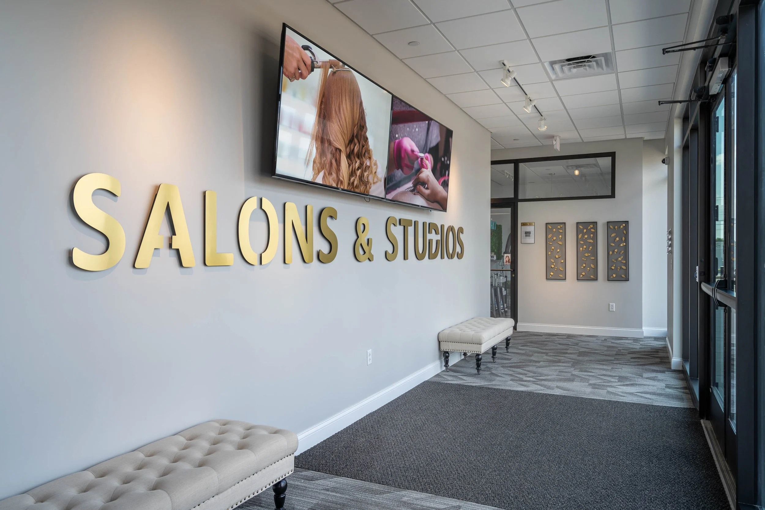 Lobby of a salon and studio with large cream-colored tufted benches, a wall with large yellow letters reading 'SALONS & STUDIOS', a wall-mounted television showing a person getting a haircut and a woman with pink hair, and framed wall art with abstra