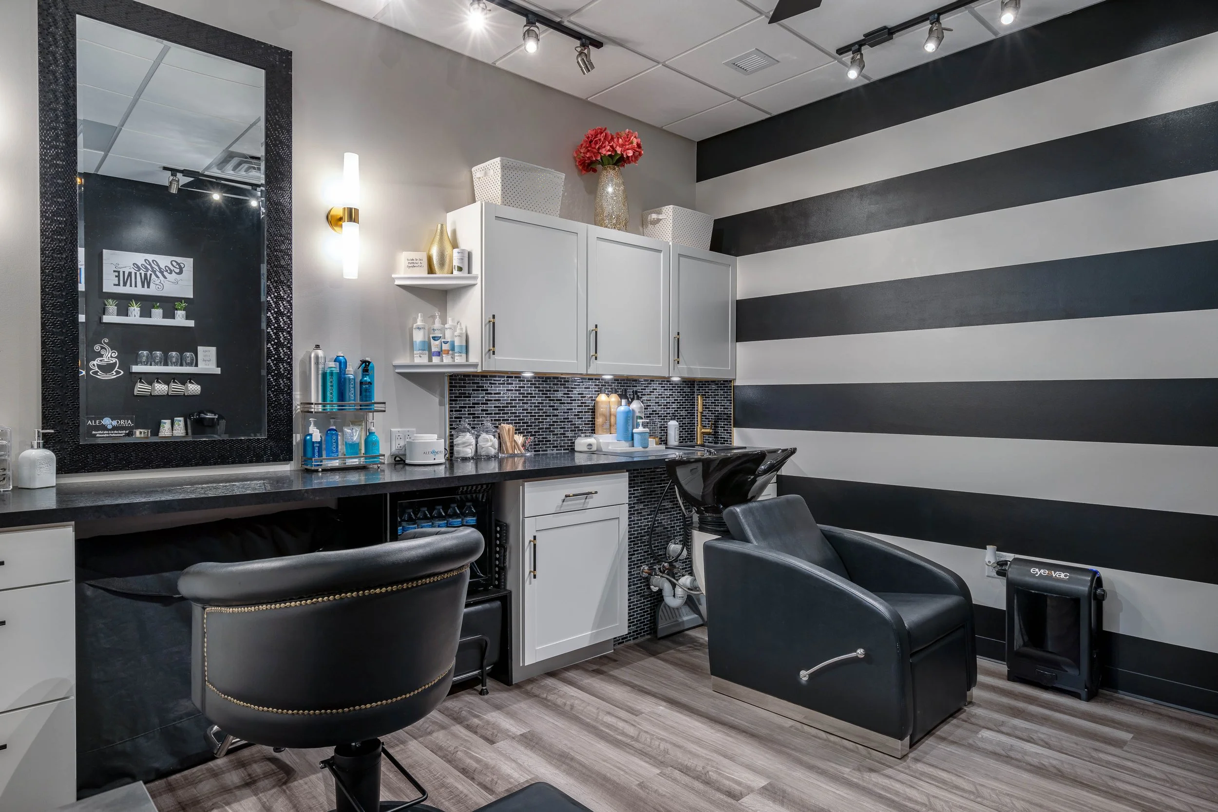 Interior of a modern hair salon with black and white striped wall, salon chairs, sink, and work station with mirror and hair products.