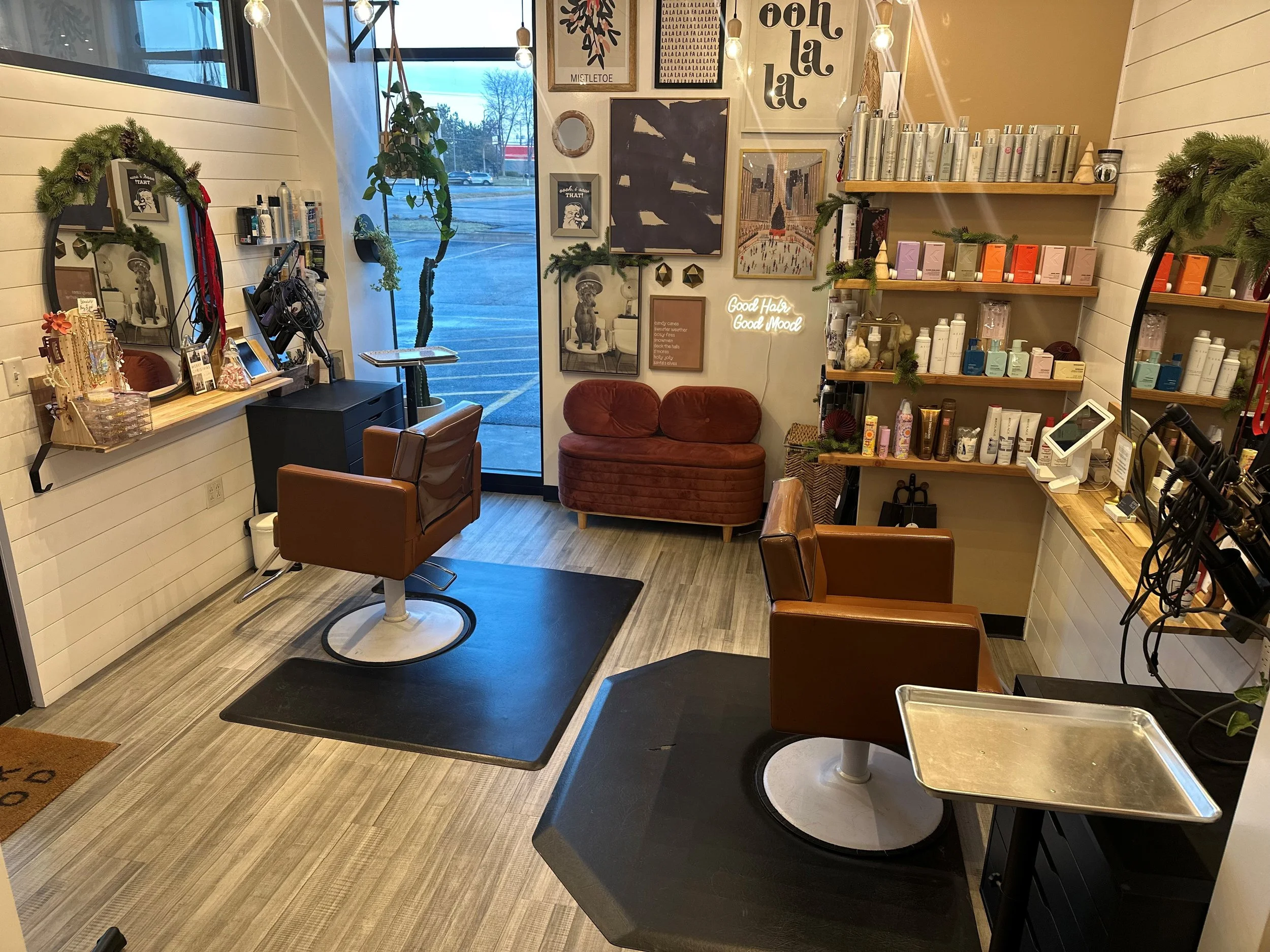 Interior of a hair salon with two brown styling chairs, shelves with hair products, a large window, and framed artwork on the walls.