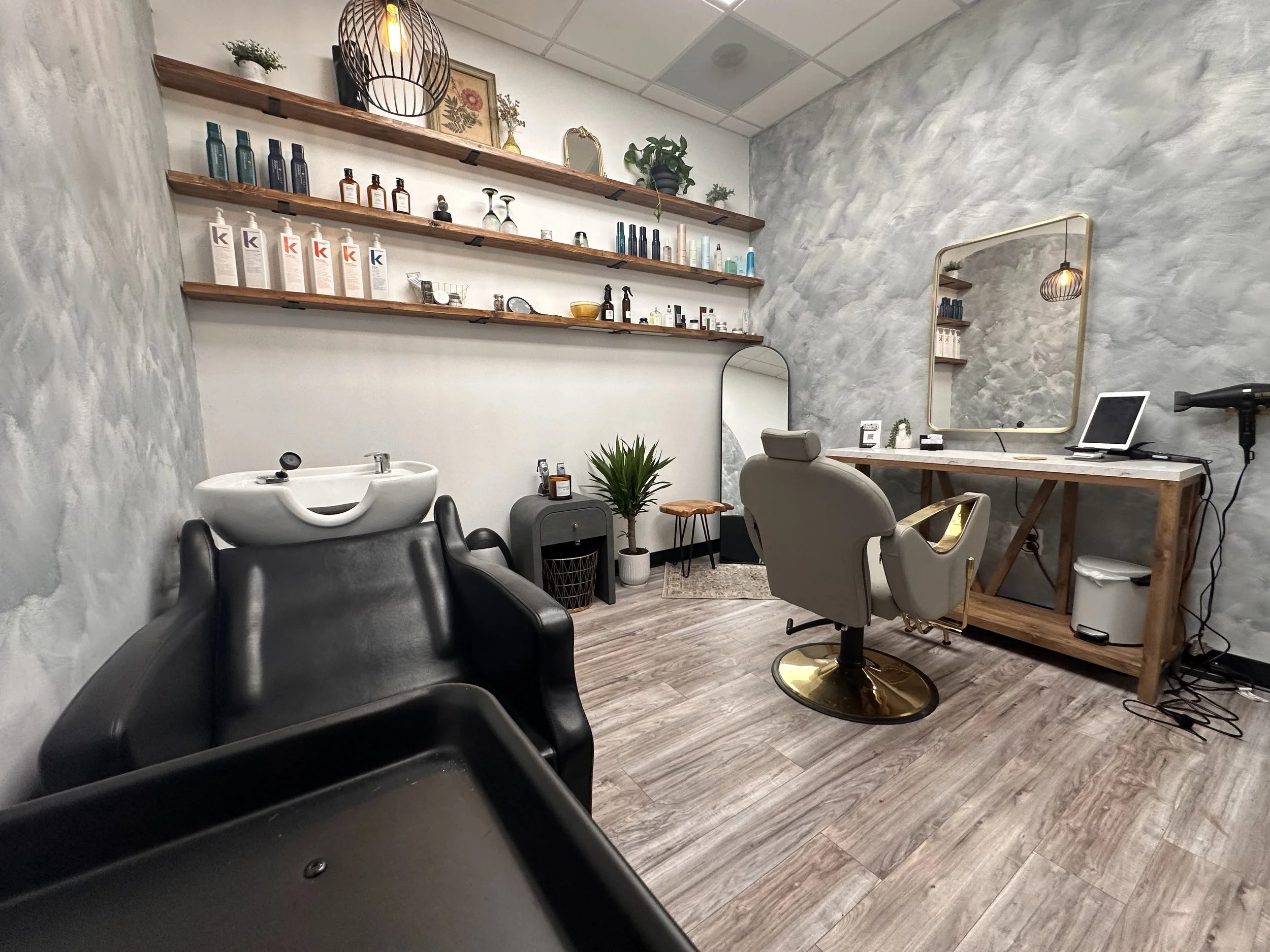 Salon with a hair washing sink, a beige salon chair, wooden shelves with beauty products, a mirror, a computer on a wooden desk, and various decorative plants and items.