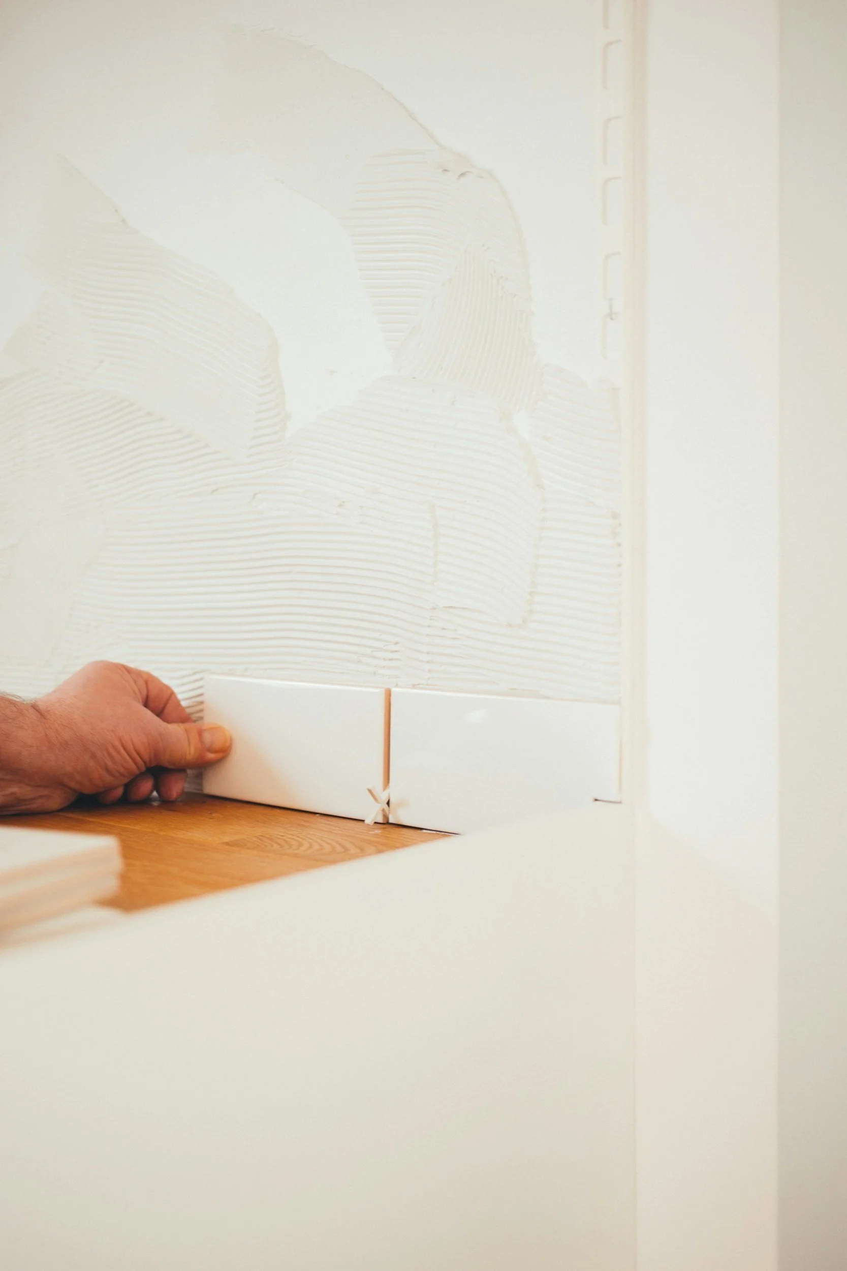 Een hand houdt een witte tegel vast terwijl deze wordt geplaatst op een muur met lijm. De muur is in aanbouw en er staat een rand van tegellijm op de vloer.