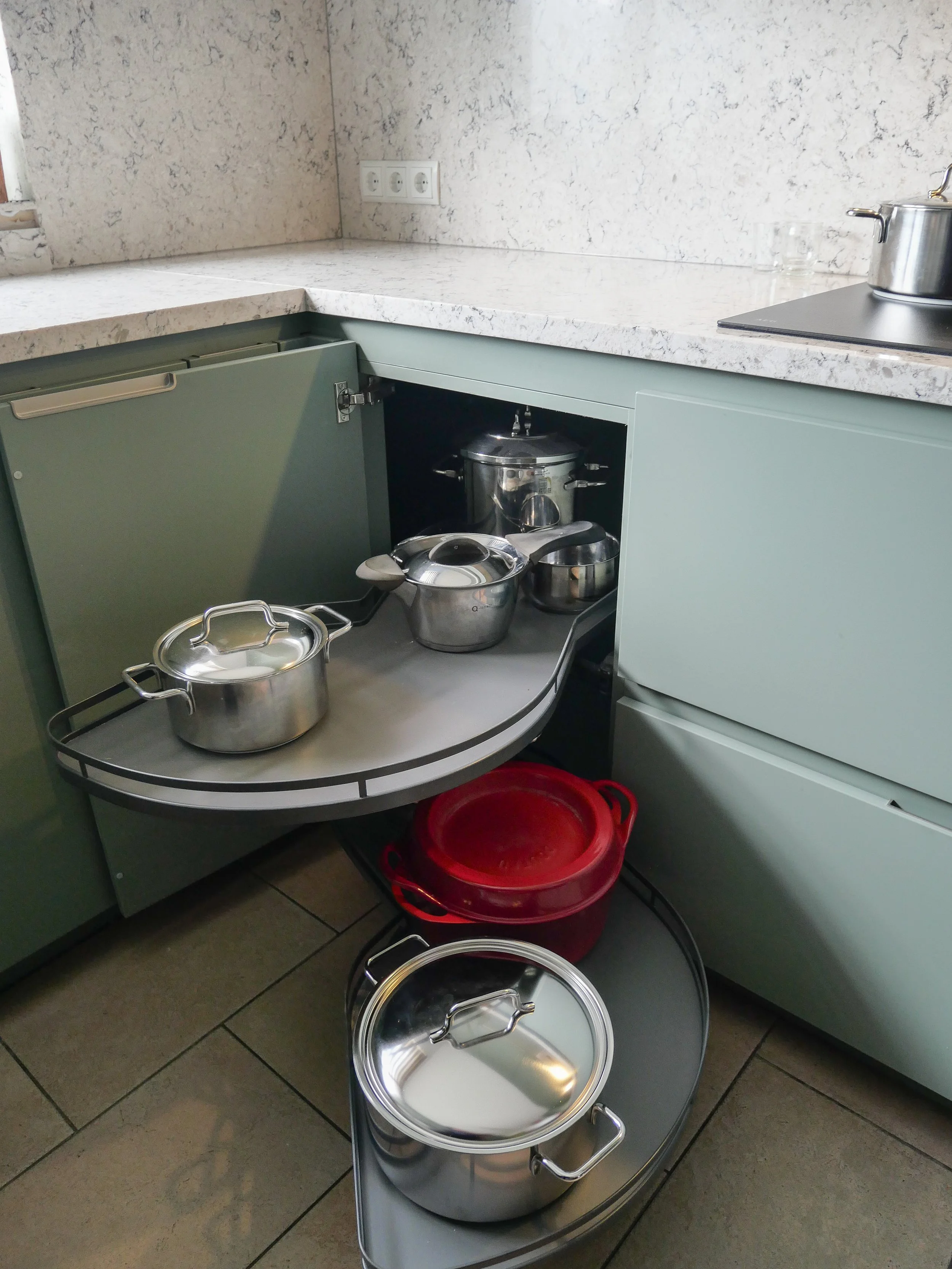 Keuken met open onderkast met verschillende potten en pannen, waaronder een ronde roestvrijstalen pan met deksel, een kleine steelpan, en een grote roestvrijstalen pan met een deksel, naast een rood onderbak en een zilveren roestvrijstalen pan op een roterende onderbak.