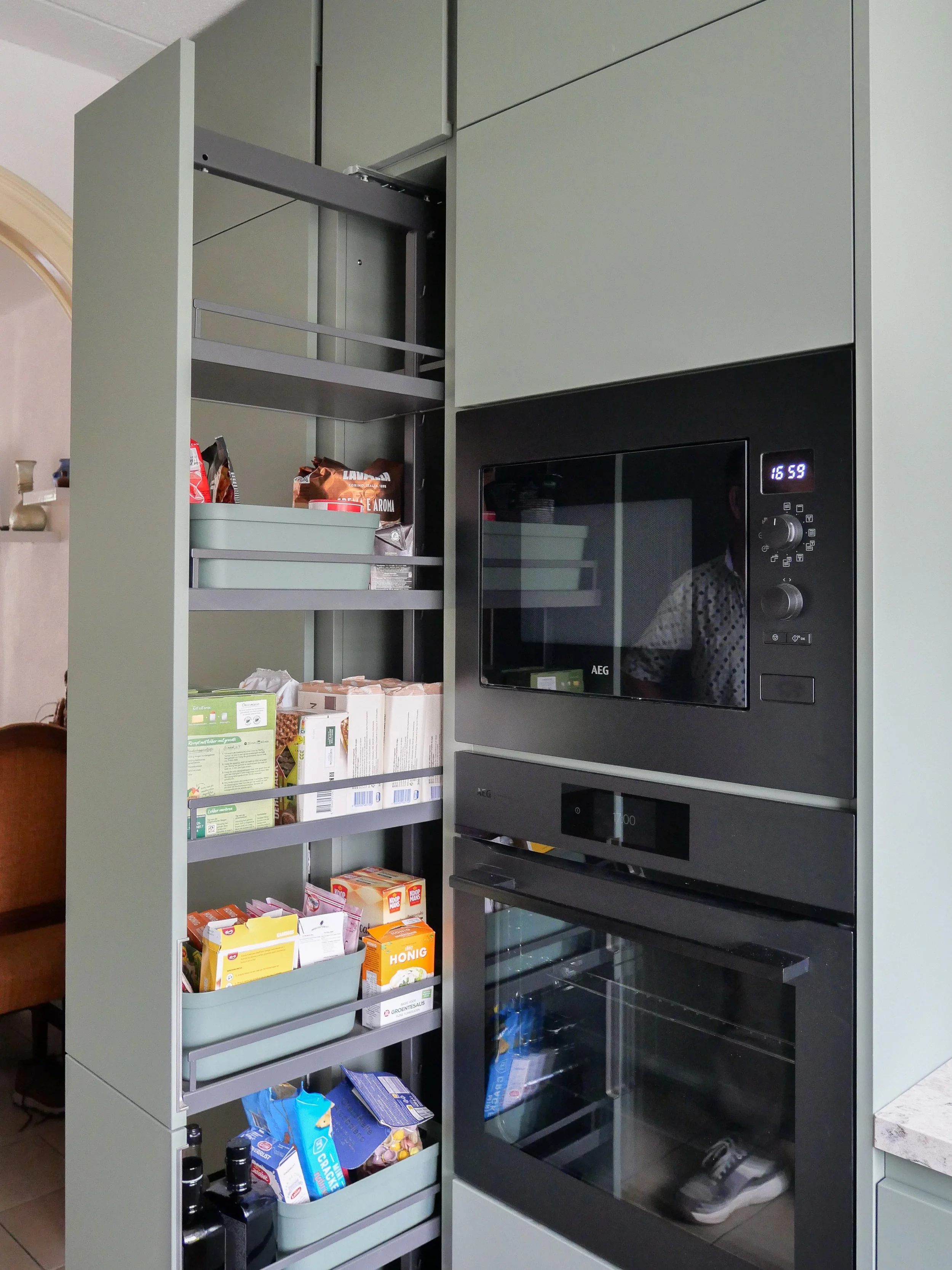 Kast met opgevulde laden, met verschillende voedingsmiddelen zoals thee, honing, en snacks, naast een ingebouwde oven en magnetron in een keuken.