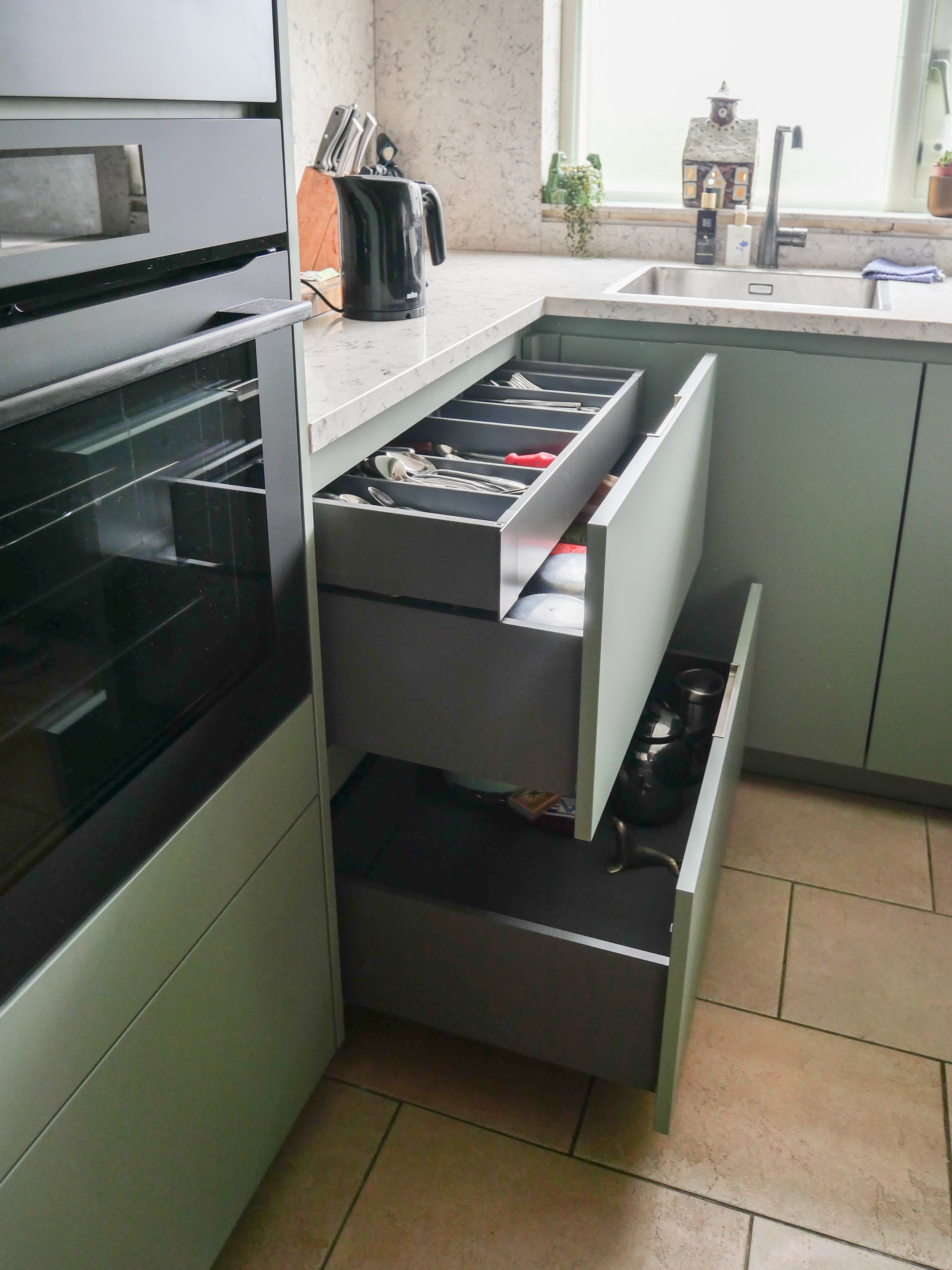 Messenkastje met bestek en enkele pannen onder de keukenkast in een moderne keuken met groene kastdeuren.