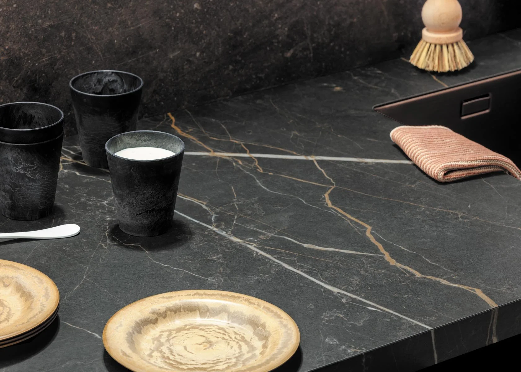 Zwarte kopjes en gouden borden op een zwarte marmeren keukenblad, met een nepbosch op de achtergrond.