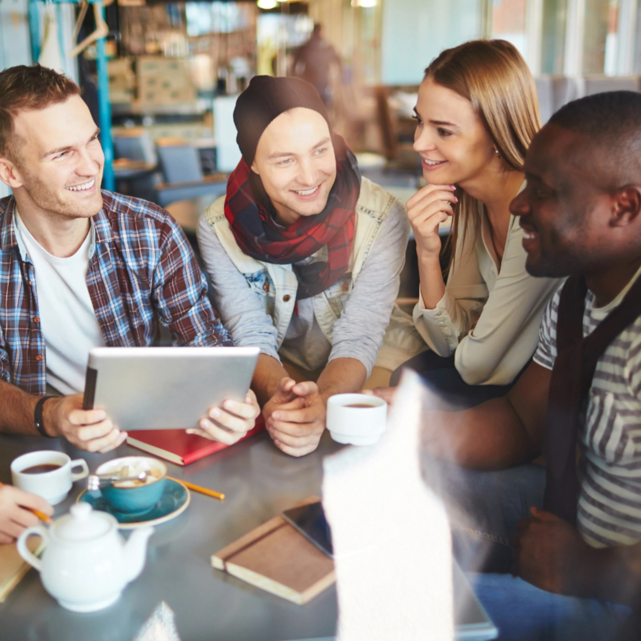 Group of friends sitting at a table in a café, smiling and conversing while holding cups of coffee and a tablet.