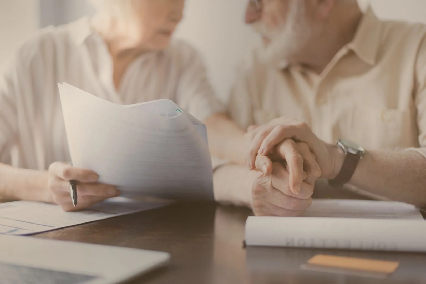An elderly man and woman holding hands across a table with documents and papers, engagement in a serious discussion.