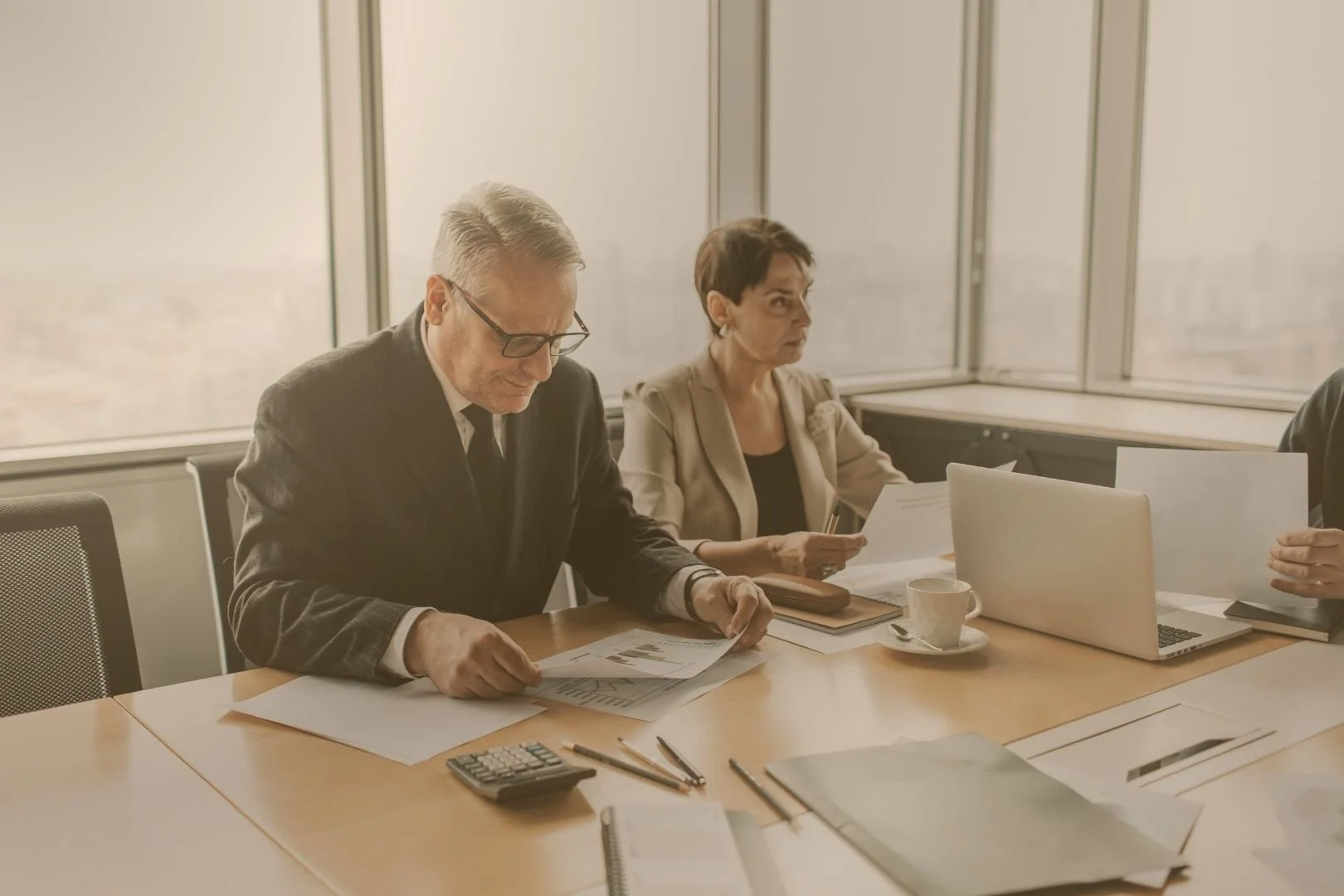Two professionals, a man and a woman, seated at a conference table with documents, a laptop, and cups, in a modern office with large windows.