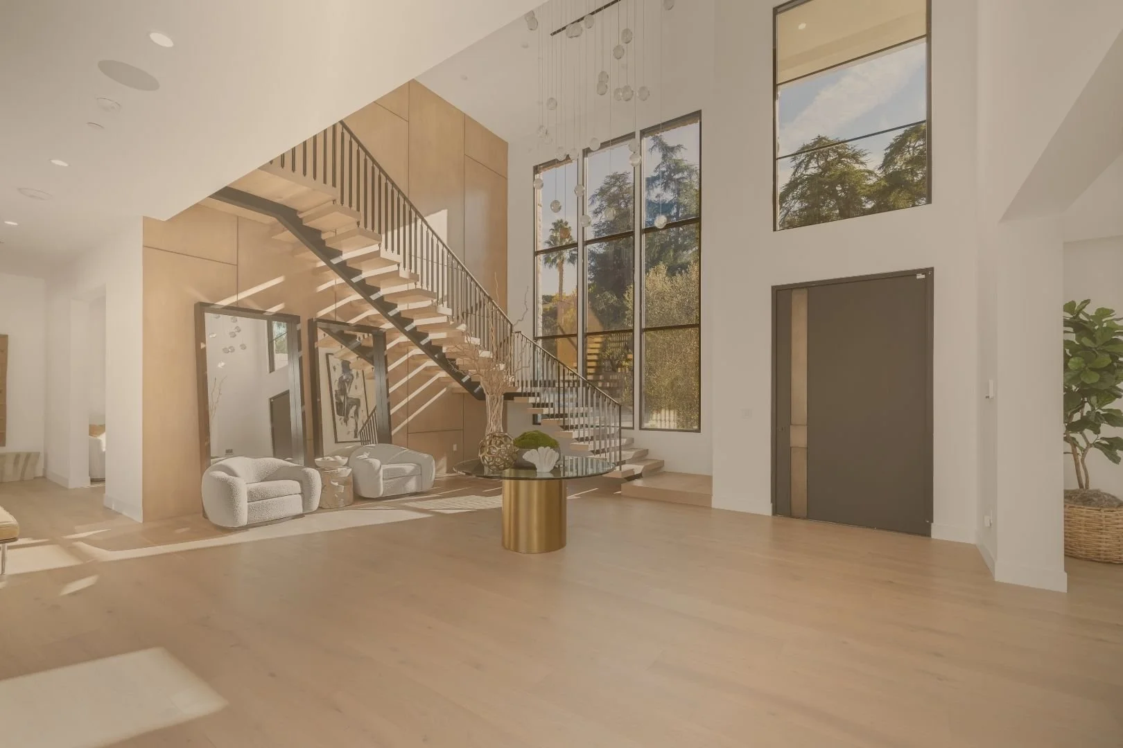 Modern living room with a high ceiling, large windows, wooden floors, and a staircase with black railing. Contains white armchairs, a gold side table, and a glass-topped table with a decorative vase and plant.