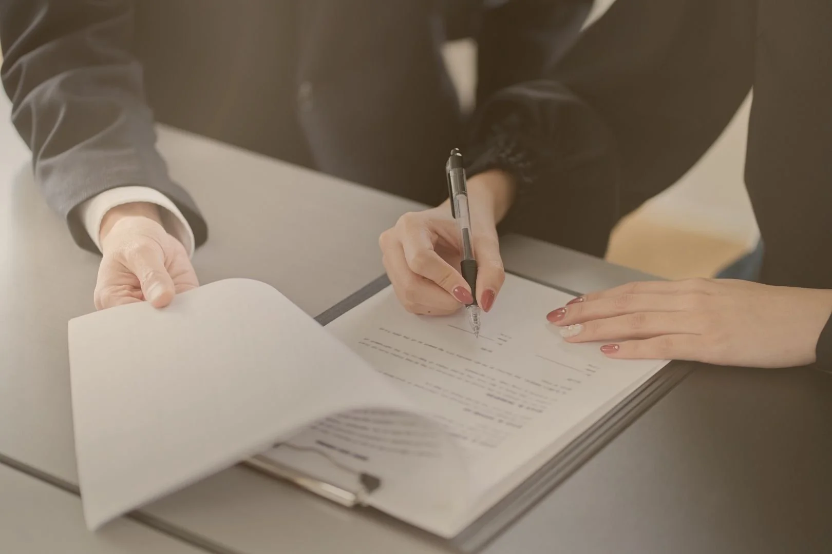 A person is signing a document on a gray table, with their right hand holding a black pen and their left hand holding the paper. The person is dressed in a dark jacket.