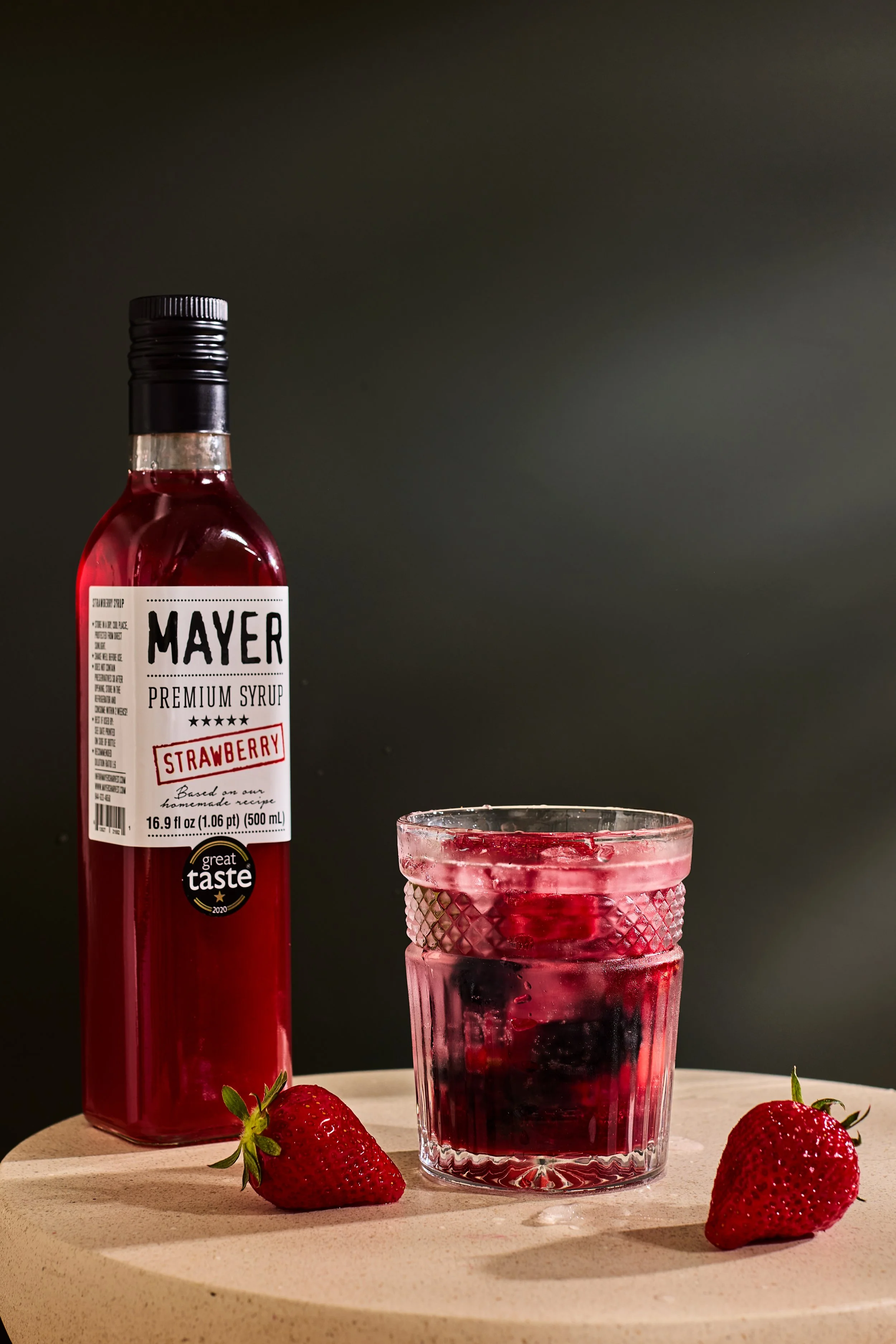 A bottle of Mayer strawberry premium syrup, a glass of strawberry syrup with ice cubes, and three fresh strawberries on a light-colored round surface.