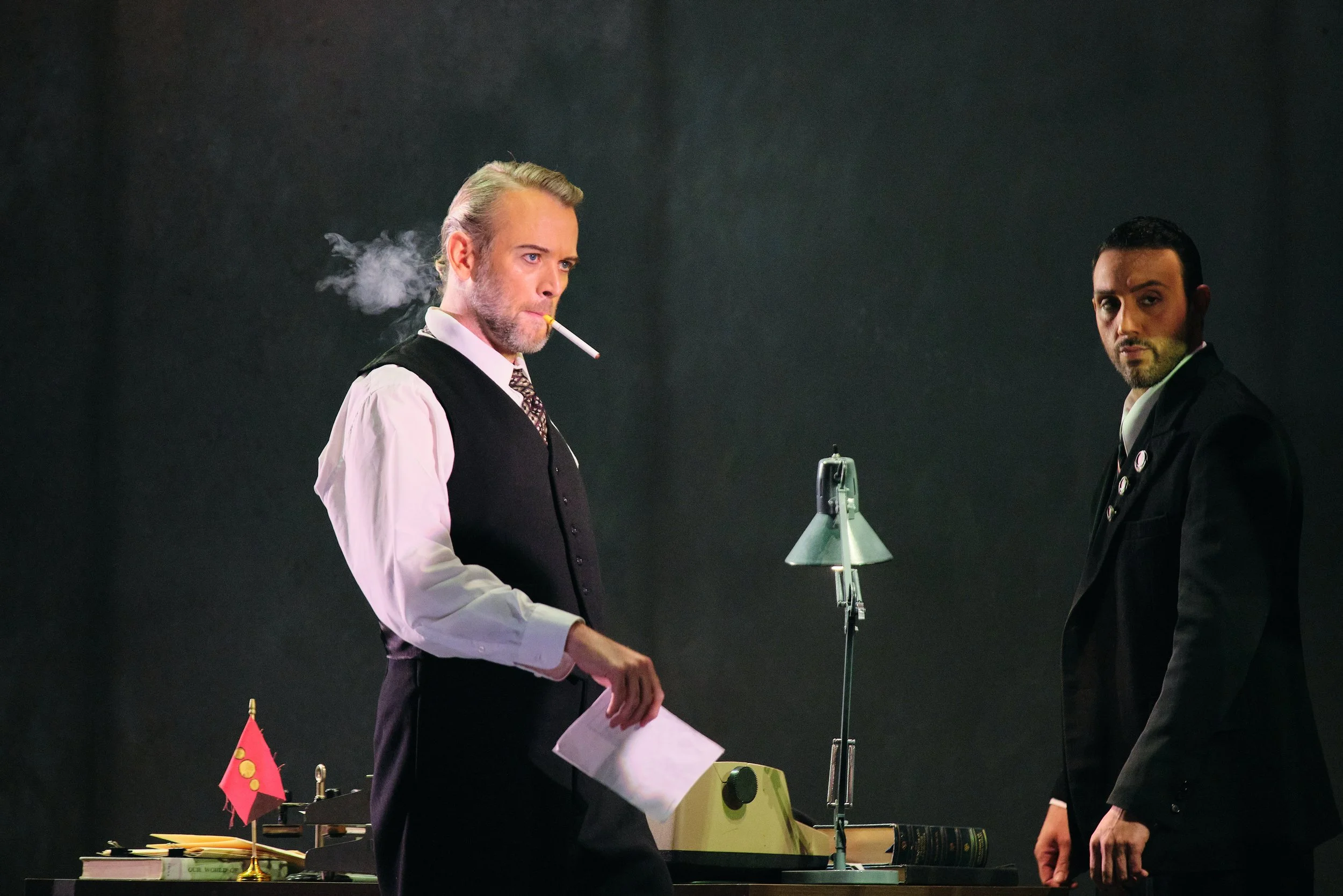 Two men dressed in formal attire, one with a cigarette in his mouth, standing near a table with flags and books, in a dark indoor setting.