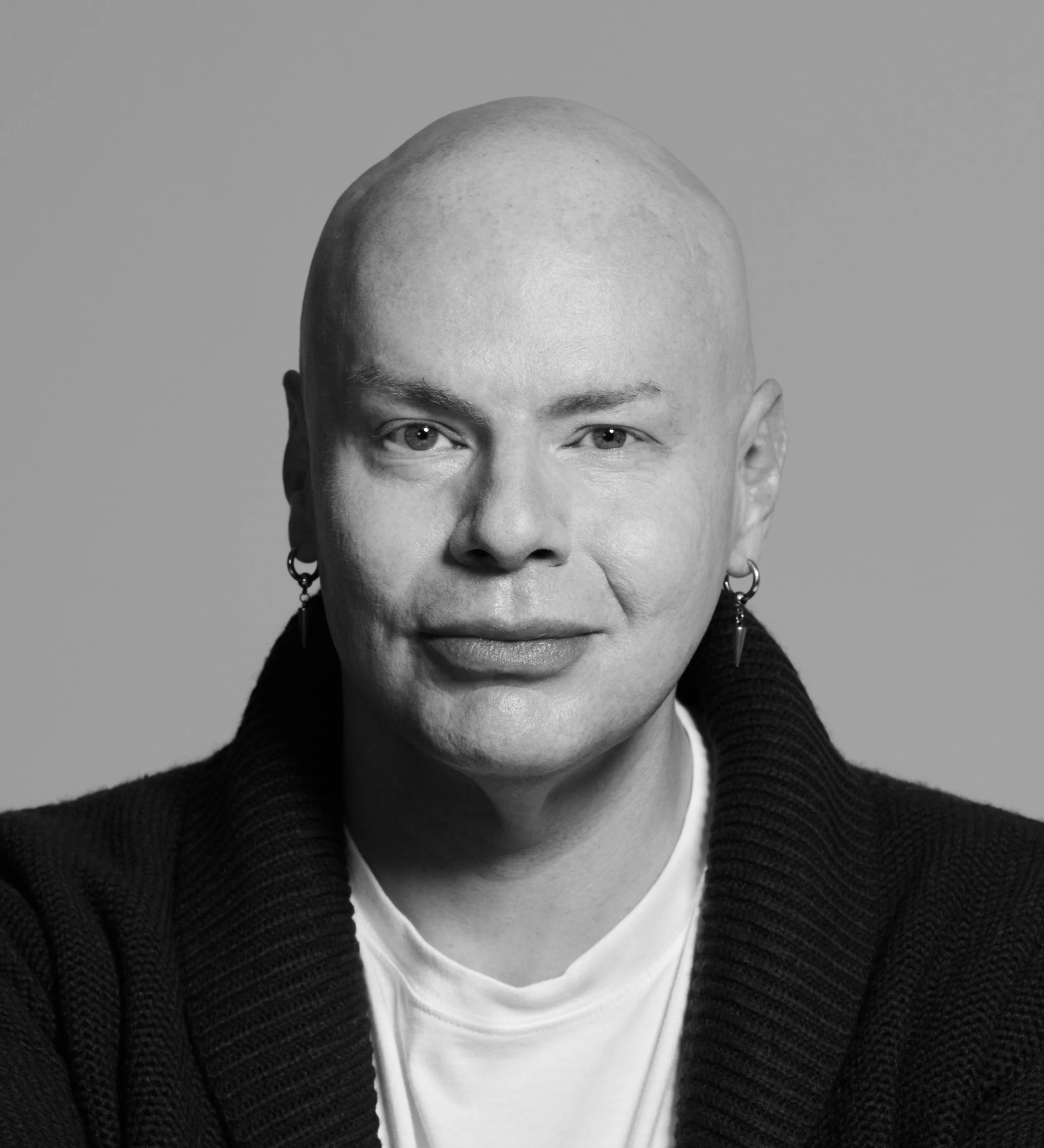 Black and white portrait of a person with a shaved head, light eyes, wearing earrings, a white shirt, and a dark knitted sweater.