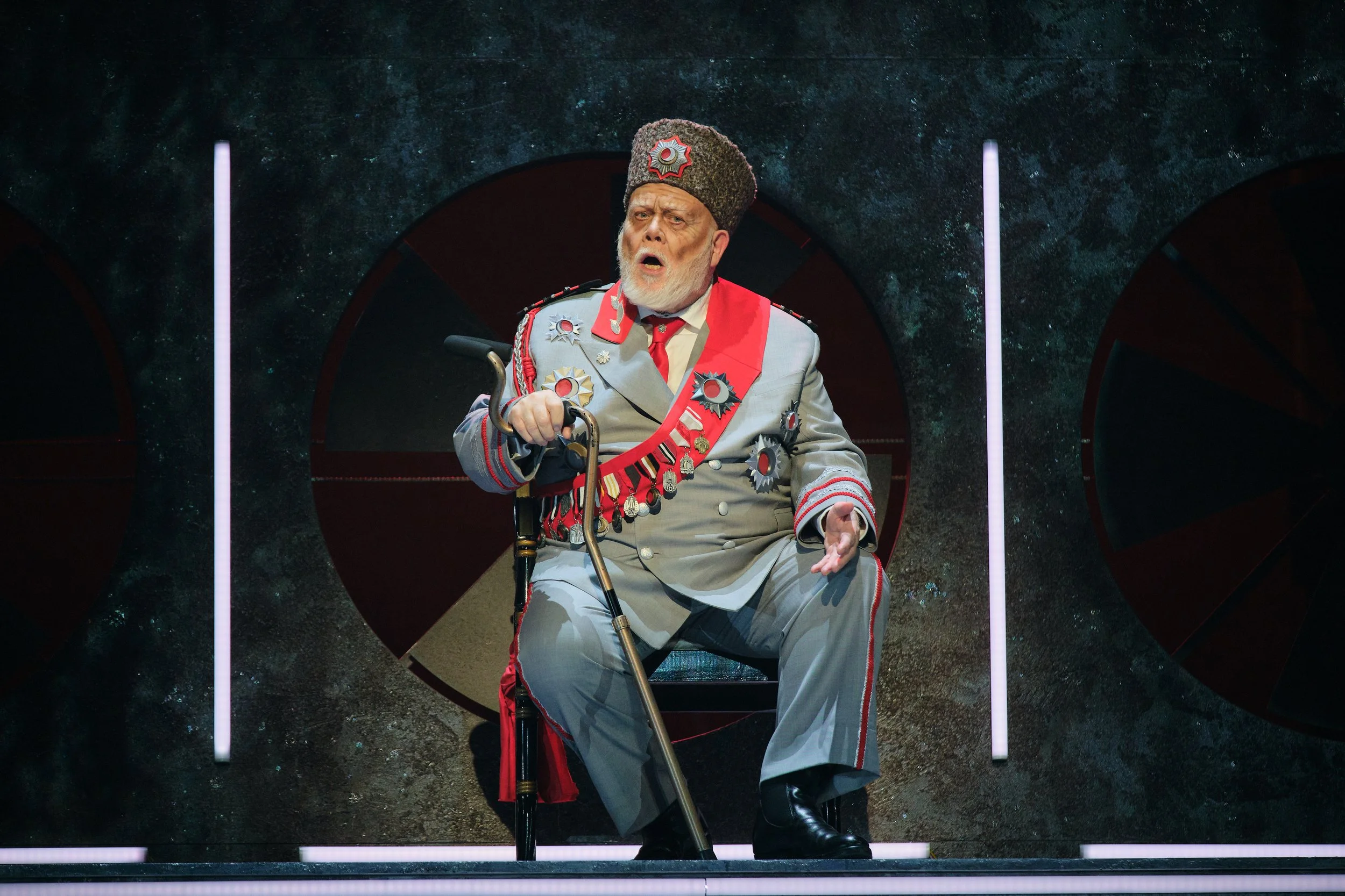 An older man dressed in a military-style uniform with medals, sitting in a chair with a cane, speaking or singing with an expressive face, on a dark stage with circular backgrounds and vertical light strips.