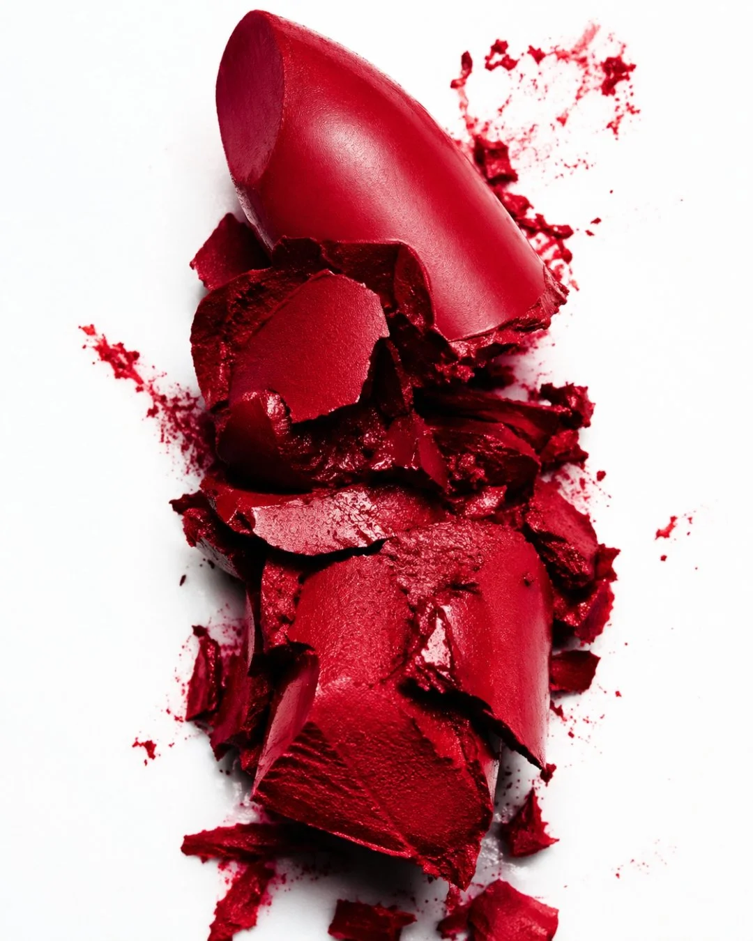 Broken red lipstick tube spilling red lipstick onto white surface.