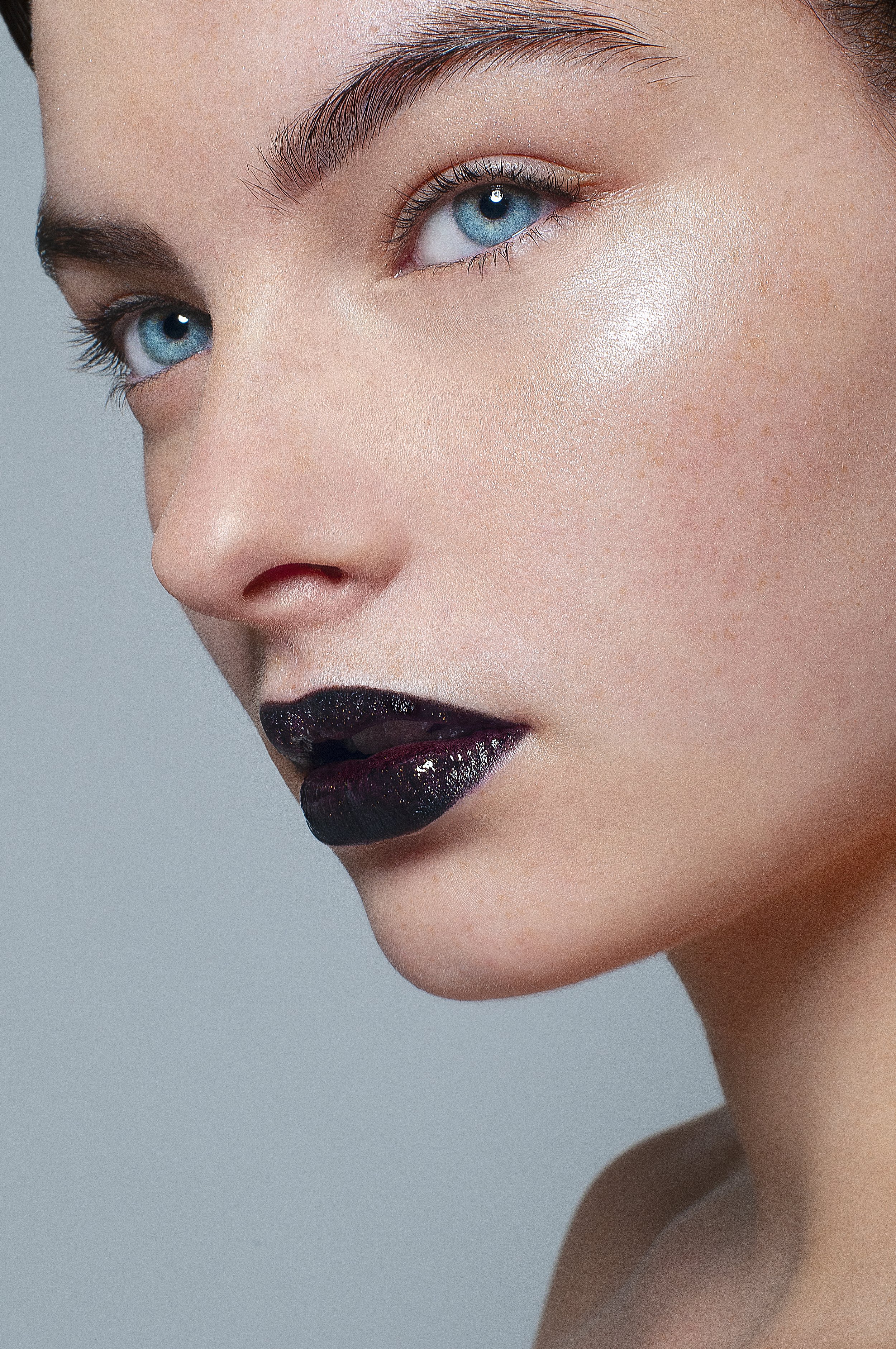 Close-up of a woman's face with bright blue eyes, dark glossy lipstick, and makeup highlighting her cheekbone.