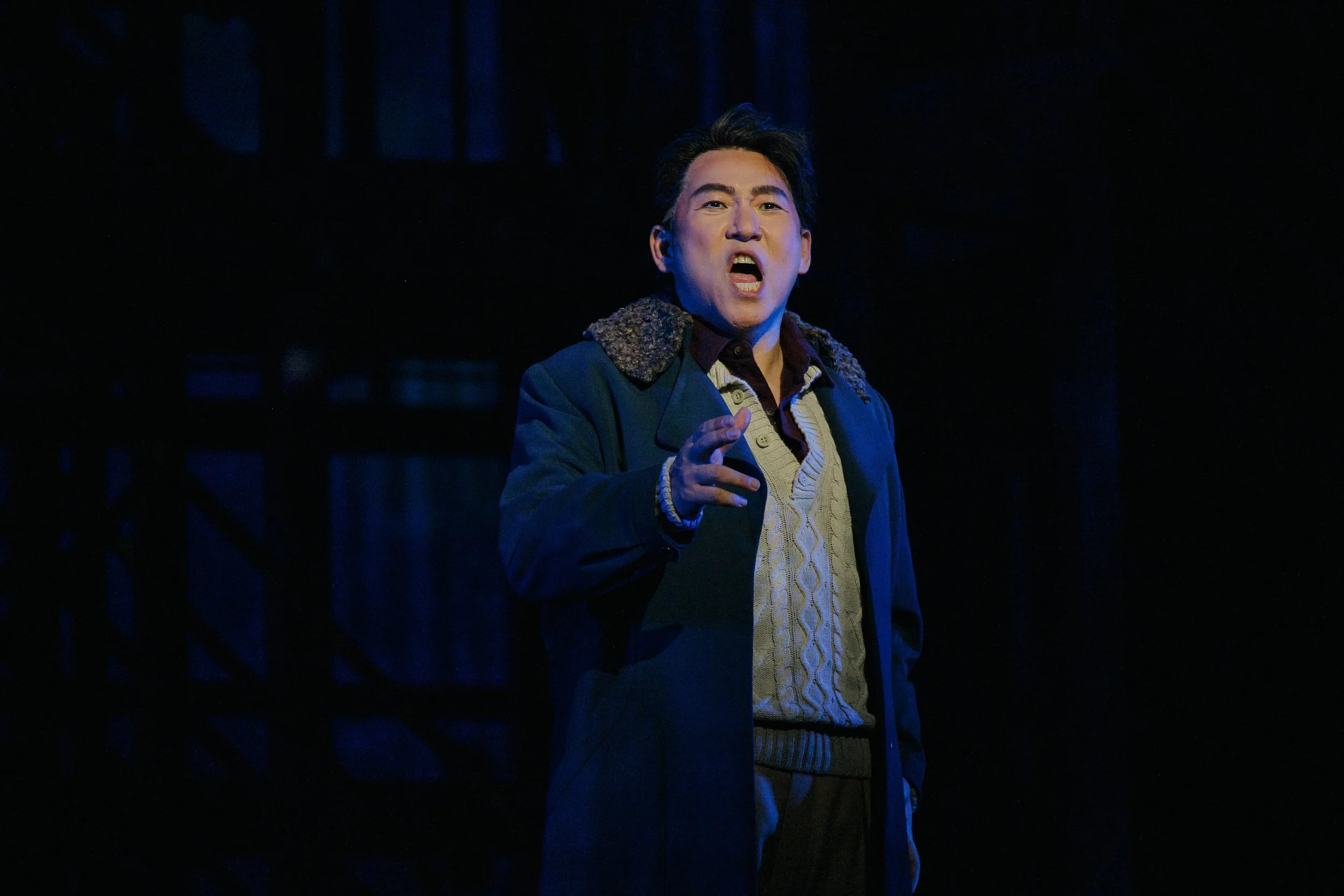 A man with dark hair speaking passionately on stage, wearing a dark coat with a fur collar, a beige sweater, and a dark shirt, against a dark background.