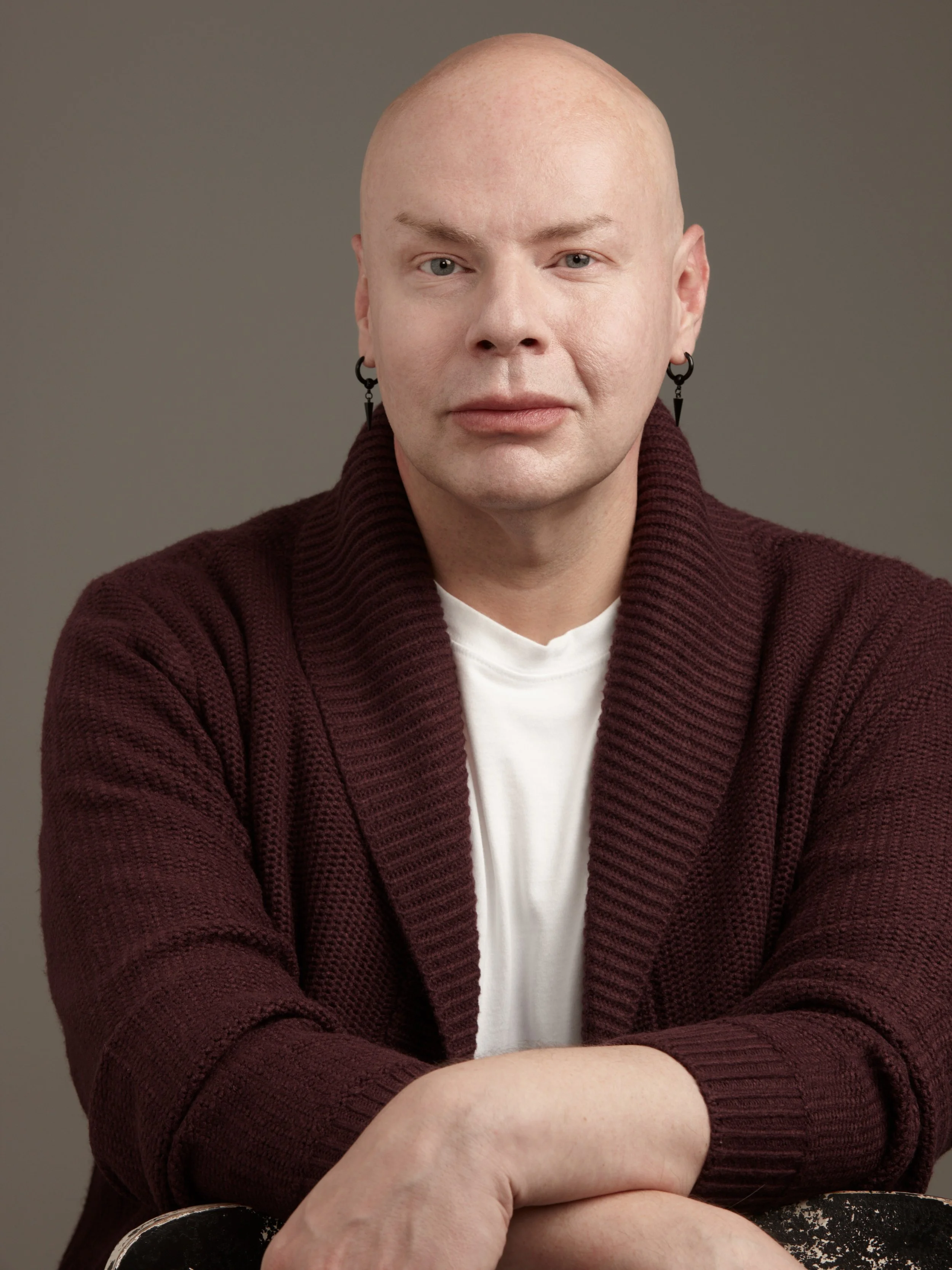 A person with light skin, a bald head, and blue eyes wearing black earrings and a burgundy knitted cardigan over a white shirt, sitting with crossed arms against a plain gray background.