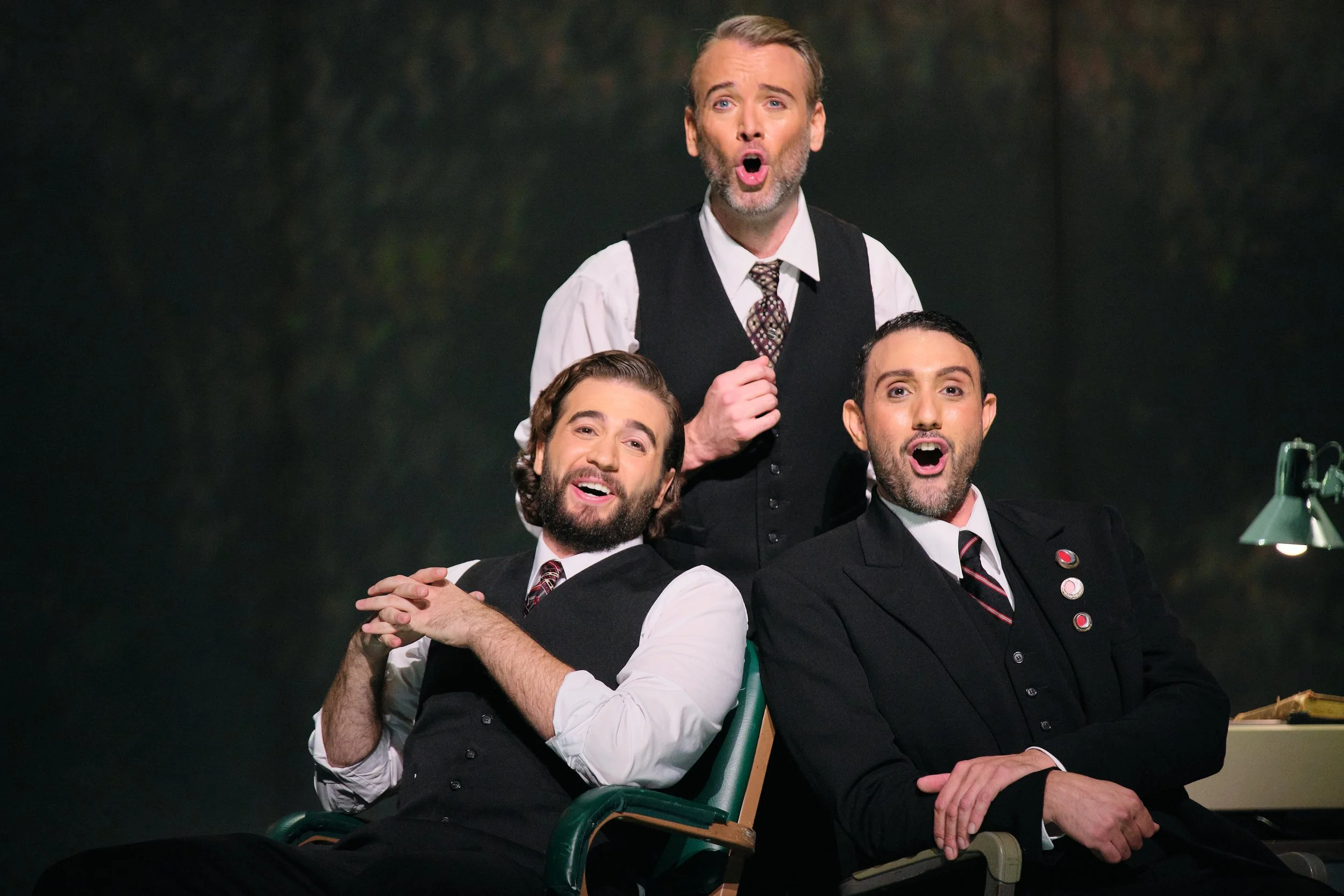 Three men in formal attire, two with beards and one with a light beard, sitting closely together in a classroom or office setting with a dark background. The man in the middle is standing and appears to be singing or speaking with an expression of surprise or excitement.