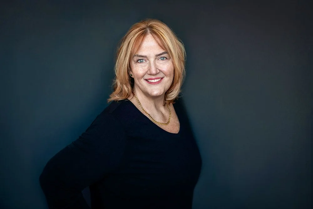 Portrait of a smiling woman with shoulder-length blonde hair, wearing a black top and a gold necklace, posing against a dark background.