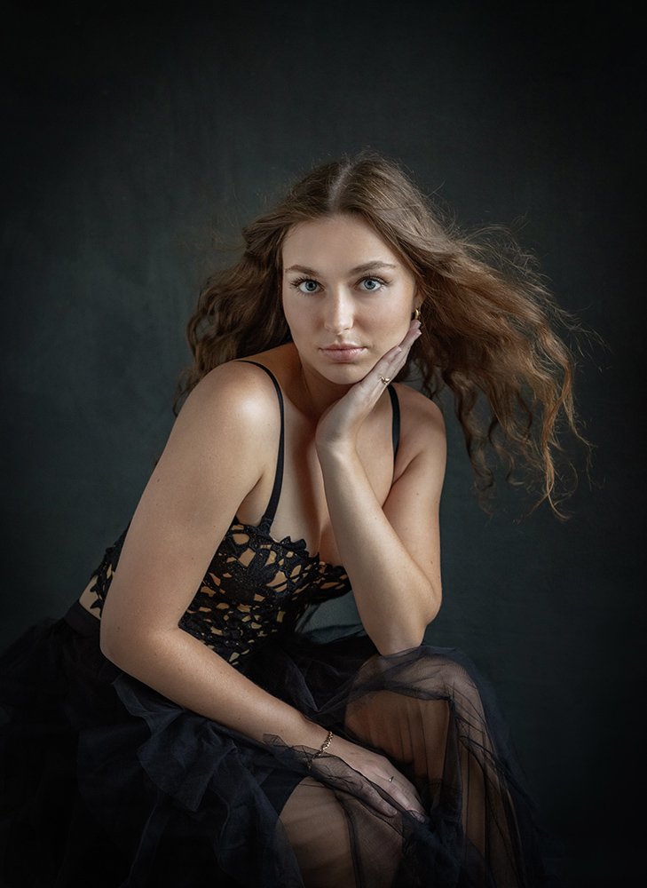 A young woman with long curly hair and blue eyes, wearing a black lace top and sheer black skirt, sitting against a dark background.