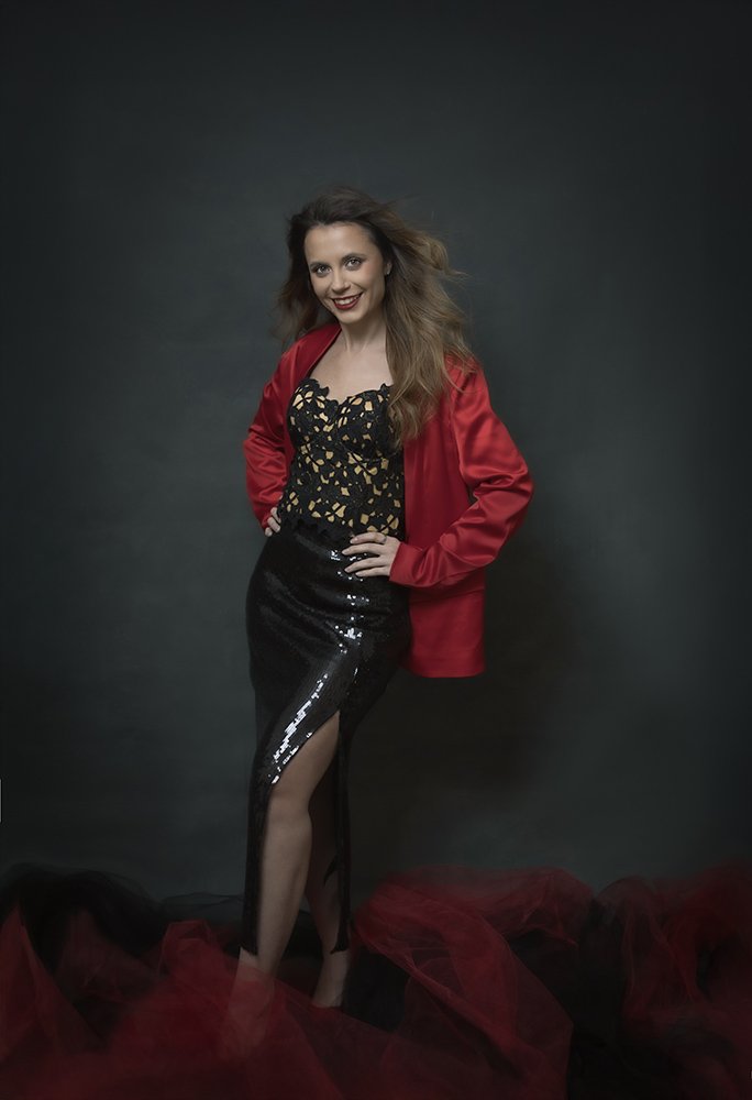 A woman with long, wavy brown hair, wearing a black dress with gold patterns, a red satin blazer, and a high-slit black sequin skirt, standing against a dark background with red tulle fabric on the floor.