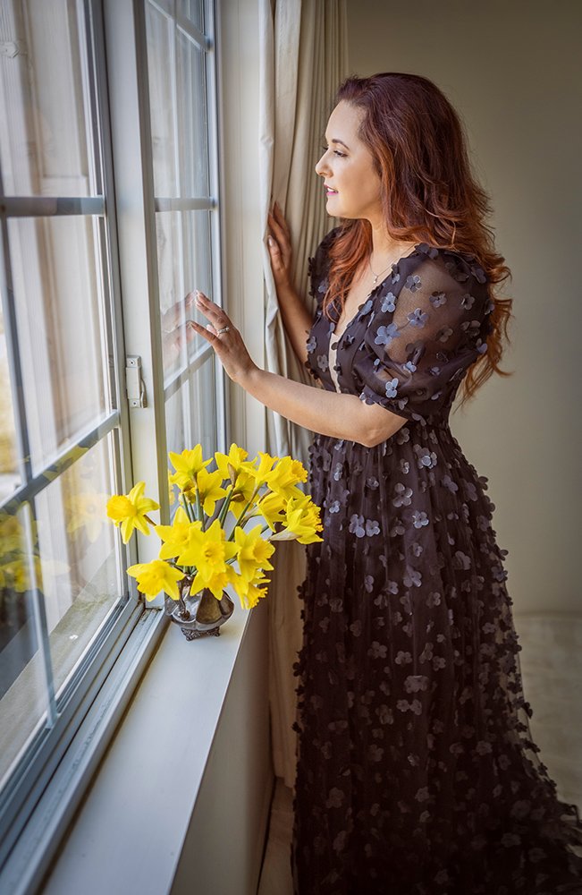 A woman with wavy, reddish-brown hair looking out a window, touching the glass with one hand, wearing a sheer black dress with purple flowers, standing next to a vase of bright yellow daffodil flowers.