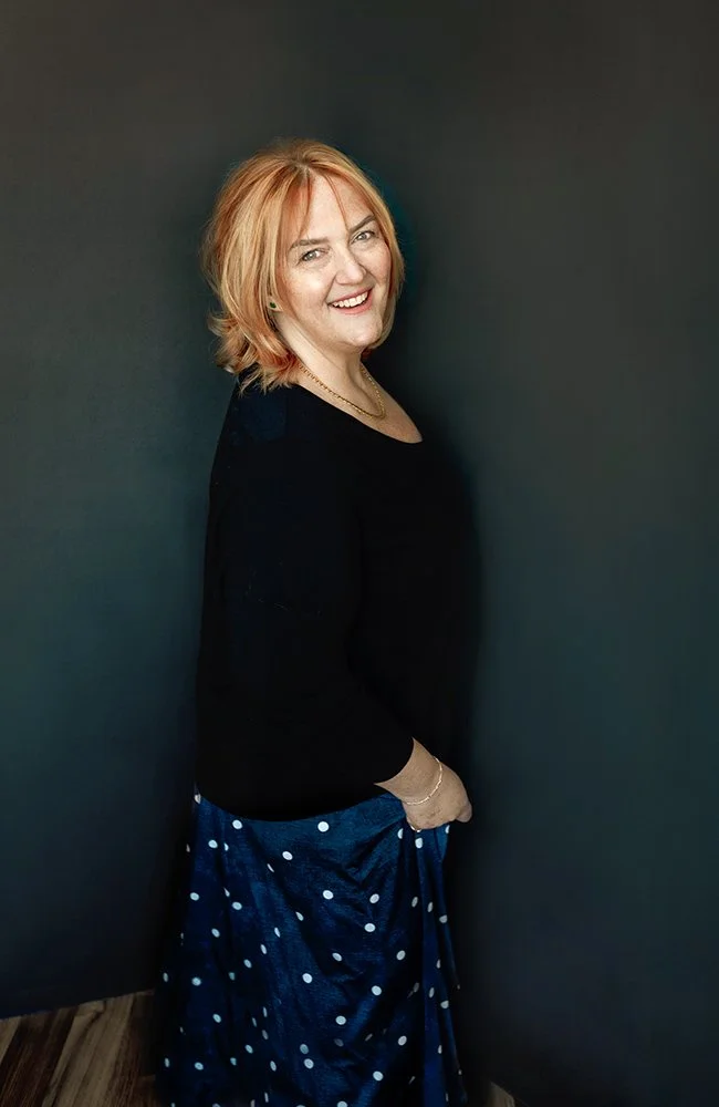 A woman with blonde hair standing against a dark wall, smiling at the camera, wearing a black top and a blue polka-dotted skirt.
