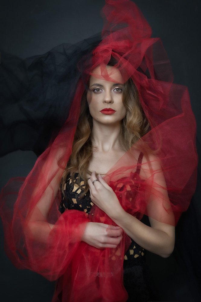 A woman with wavy blonde hair and red lipstick, wearing black lingerie, is surrounded by sheer red and black fabric against a dark background.