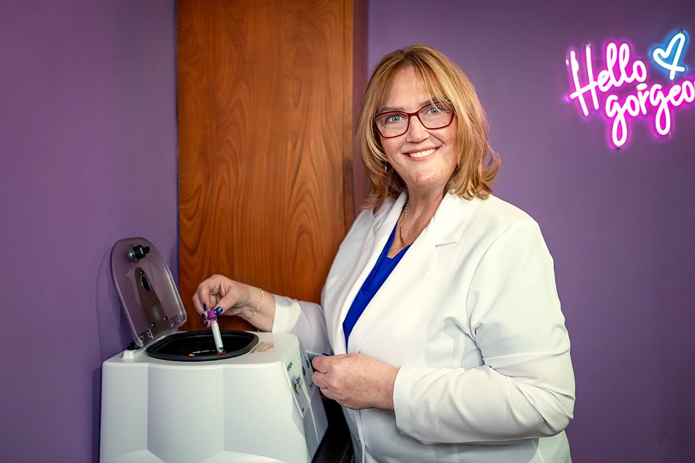 A smiling woman with glasses and blonde hair in a white lab coat using a small centrifuge in a purple-walled room with a neon sign that says "Hello Gorgeous" in pink and blue.