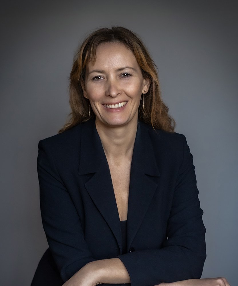 A woman with shoulder-length light brown hair smiling, wearing a dark blazer against a gray background.