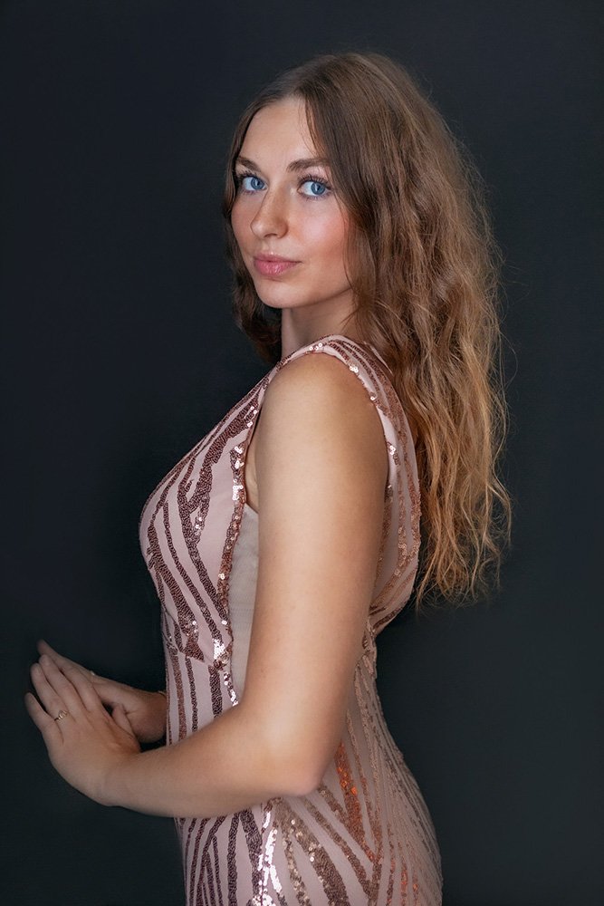 A woman with wavy, reddish-brown hair and blue eyes, wearing a sleeveless, pink, and rose gold sequined dress with vertical stripes, standing against a dark background.