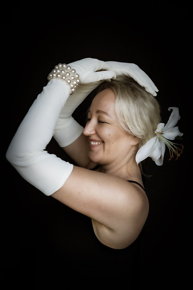 A woman with short blonde hair and a white lily in her hair, smiling with eyes closed, wearing long white gloves with pearl bracelets, posed against a black background.