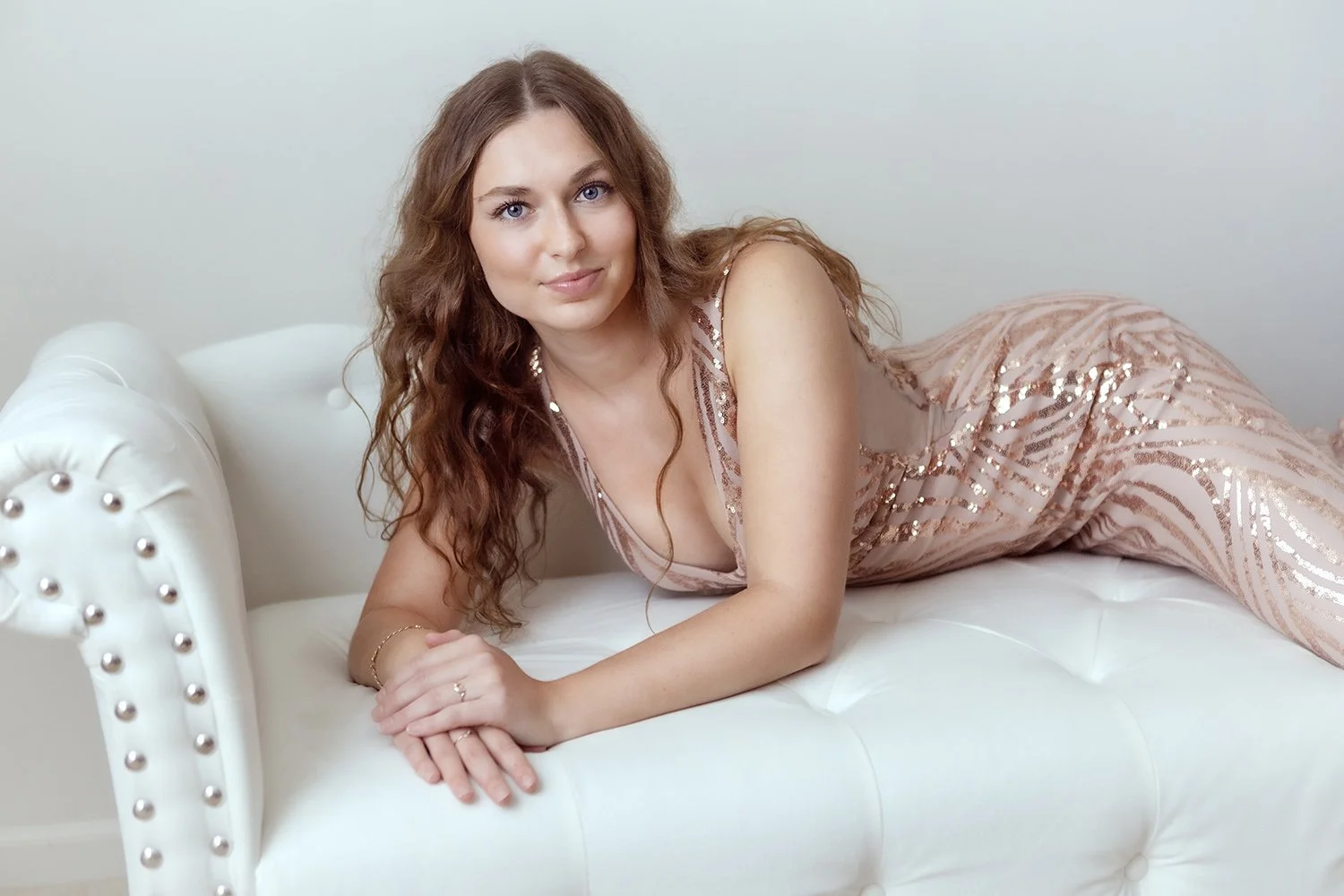 A woman with long wavy brown hair, blue eyes, wearing a pink sequin dress, lying on a white tufted chaise lounge.