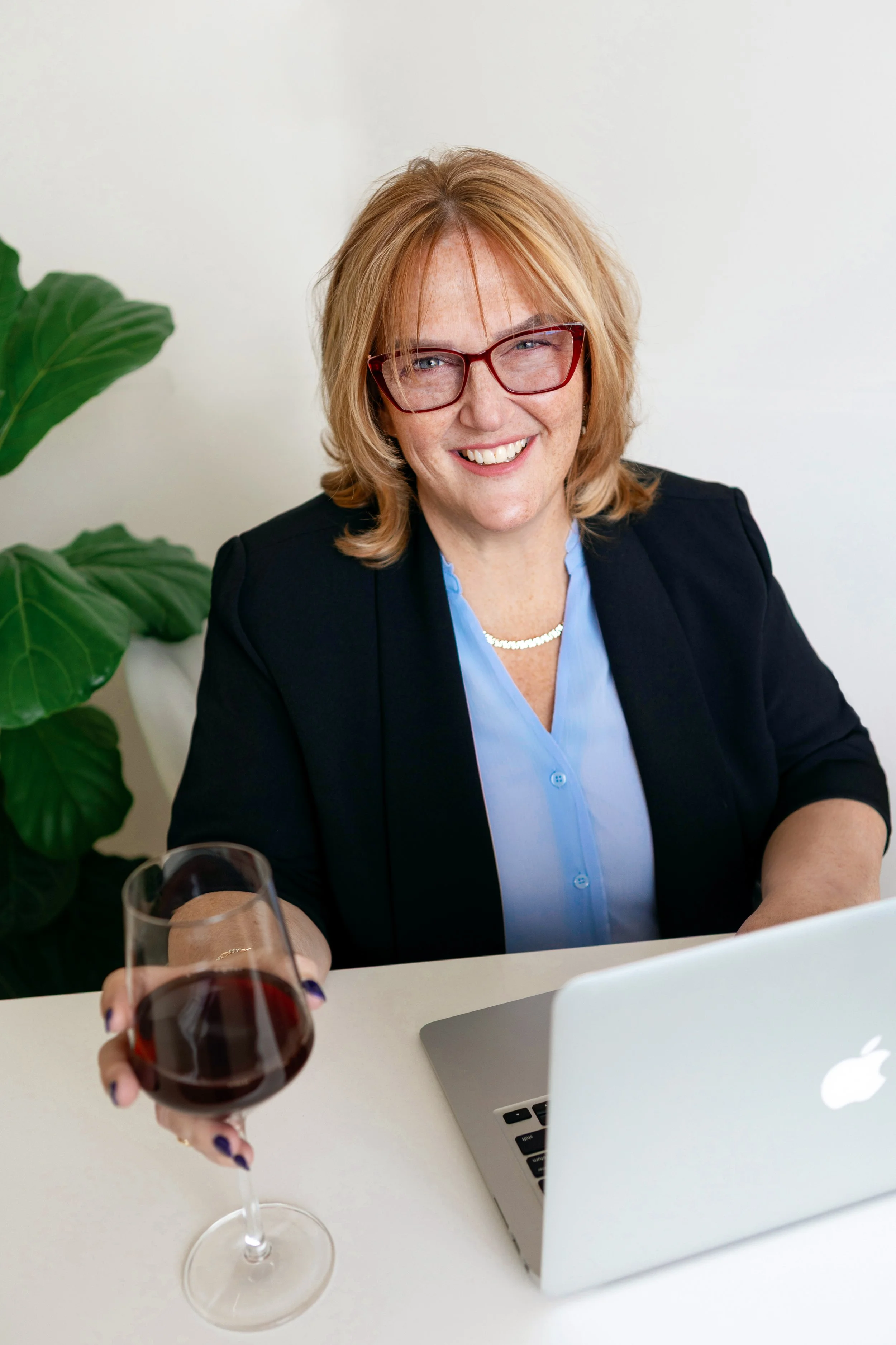 Smiling woman with red glasses holding a glass of red wine, sitting at a desk with a laptop, in front of green plant.