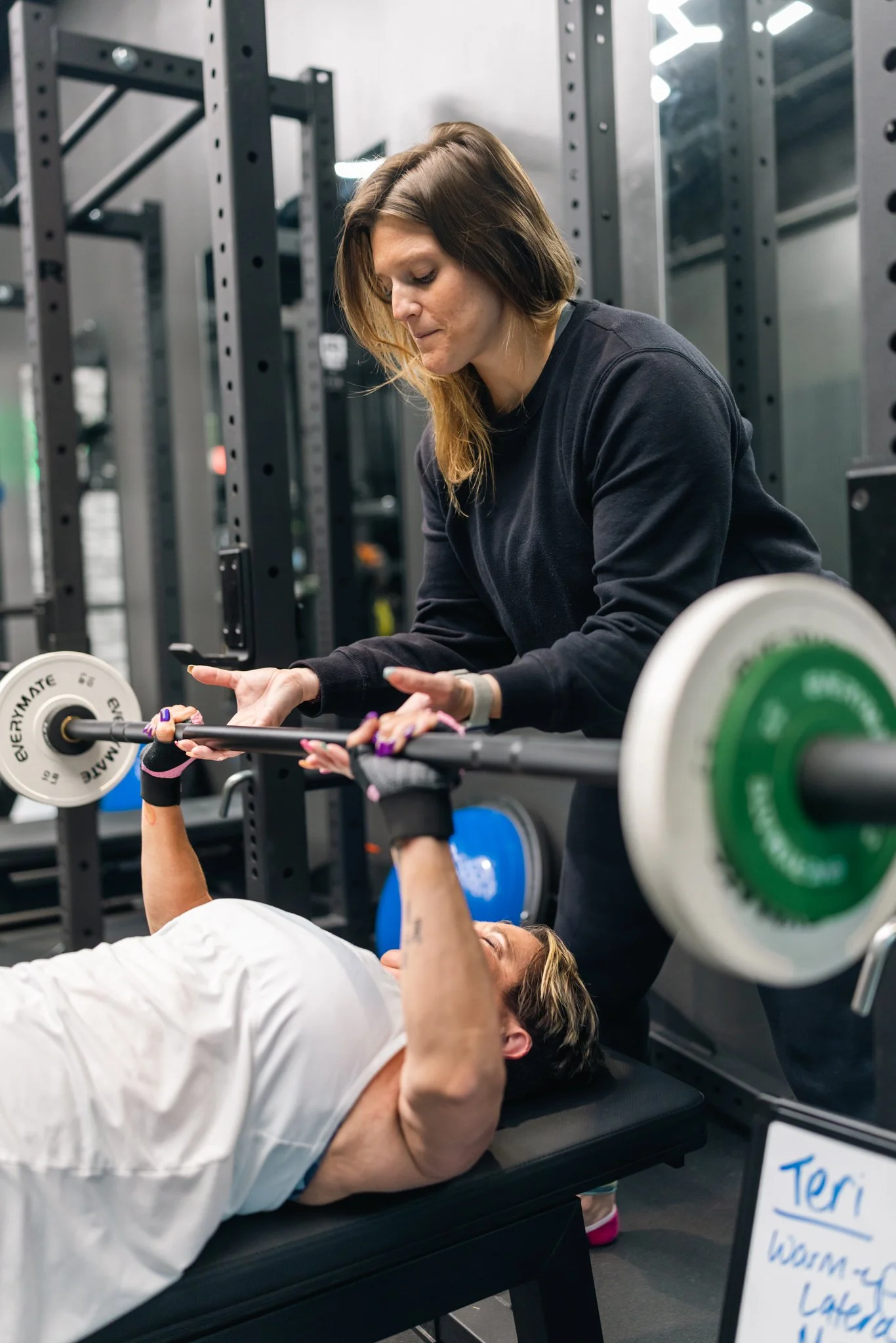 A woman spotting a man lifting weights at the gym, with weightlifting equipment and a sign labeled 'Teri' in the background.