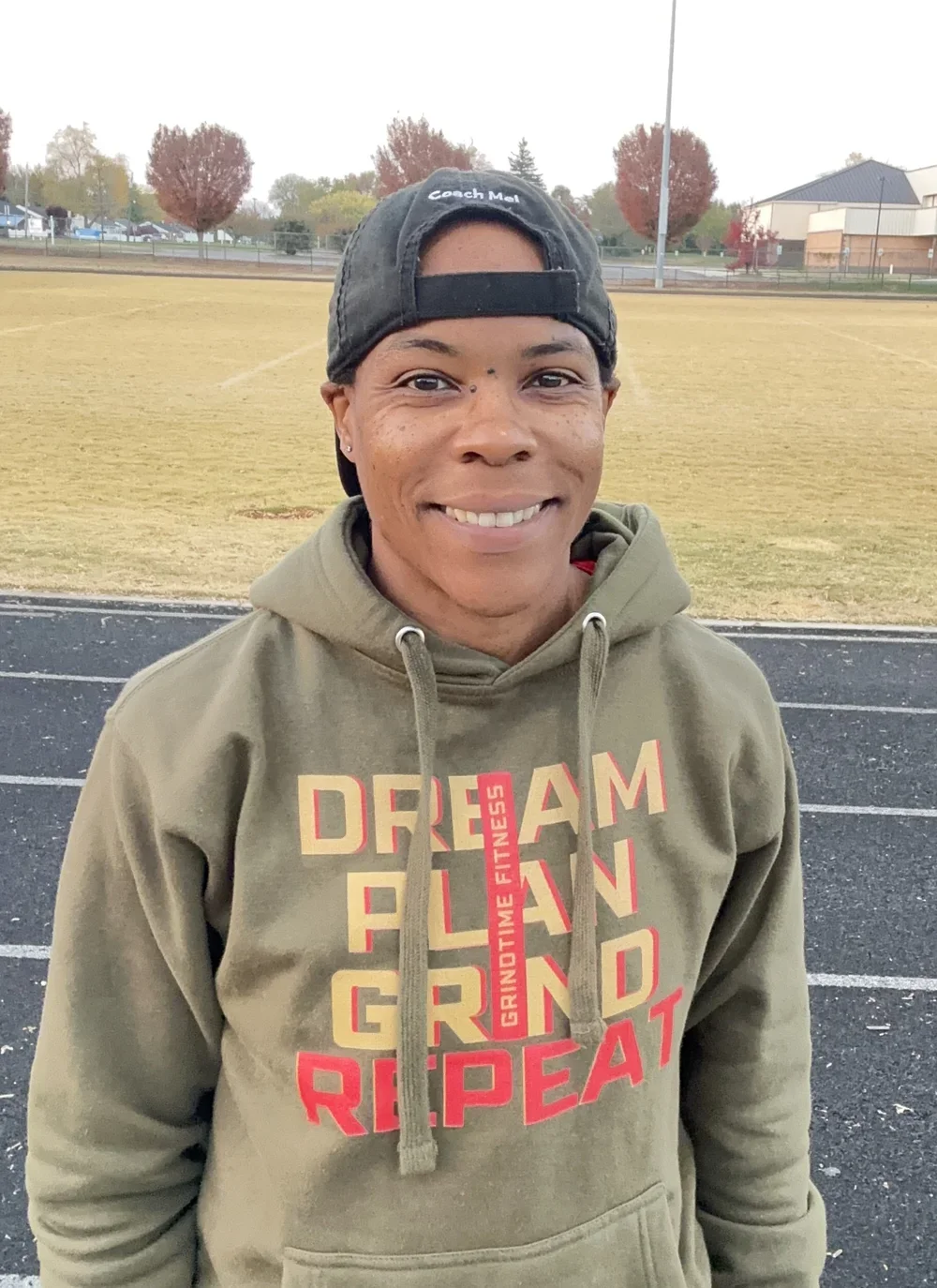 A smiling woman wearing a backwards black cap and a green hoodie with red and yellow text standing on a running track. The background includes a grassy field, trees with autumn foliage, and a building.