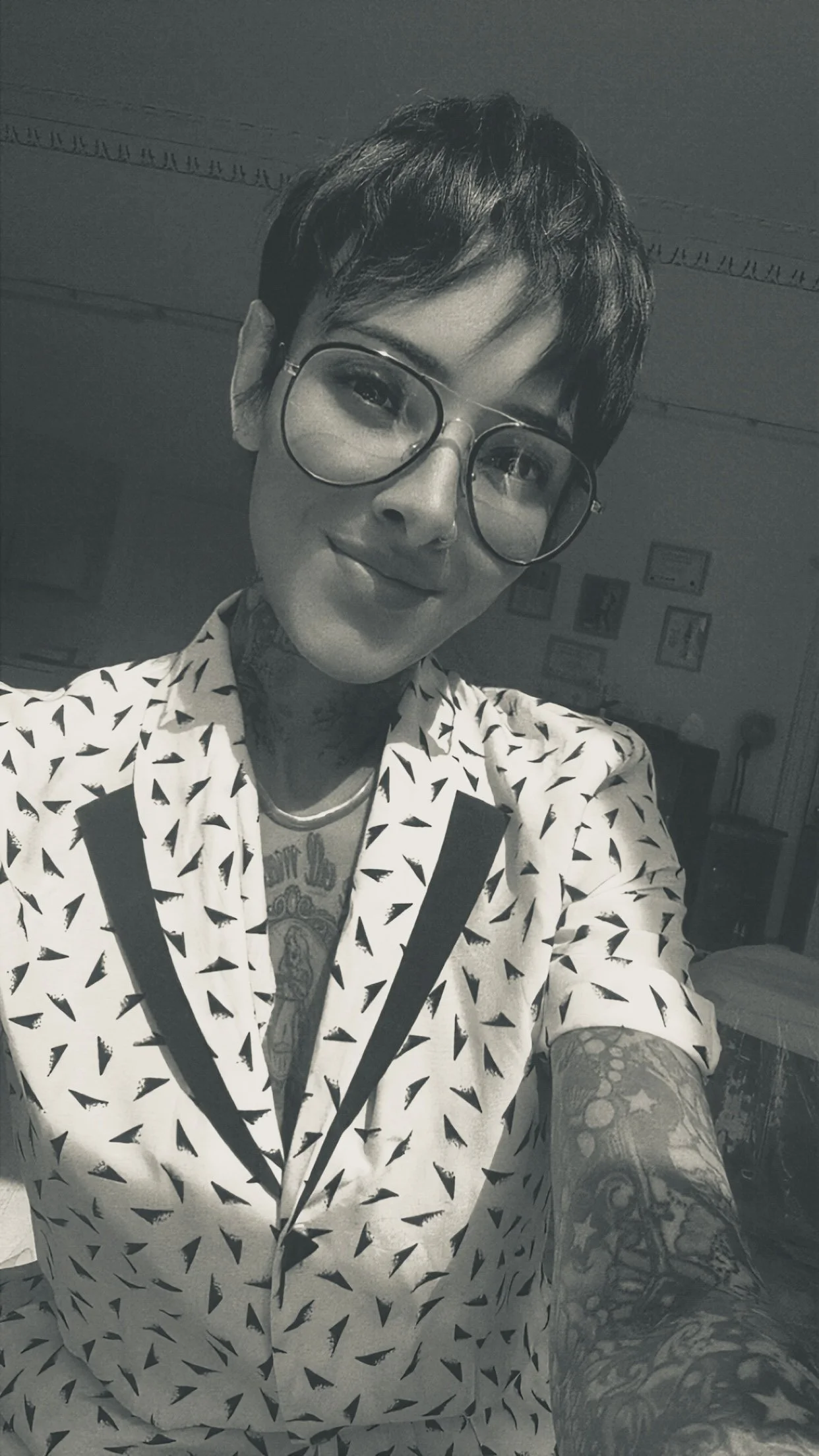 A person with short dark hair, wearing glasses and a shirt with a black triangle pattern on a white background, is taking a selfie in a room with framed pictures on the wall and a bookshelf in the background. They have tattoos visible on their neck and arm.