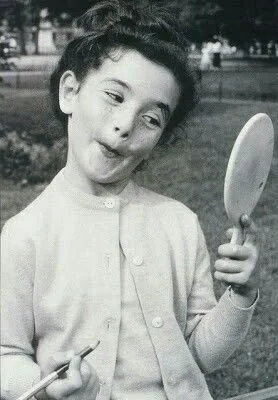 A young girl with short hair looking into a mirror while holding a comb in one hand outside in a park.