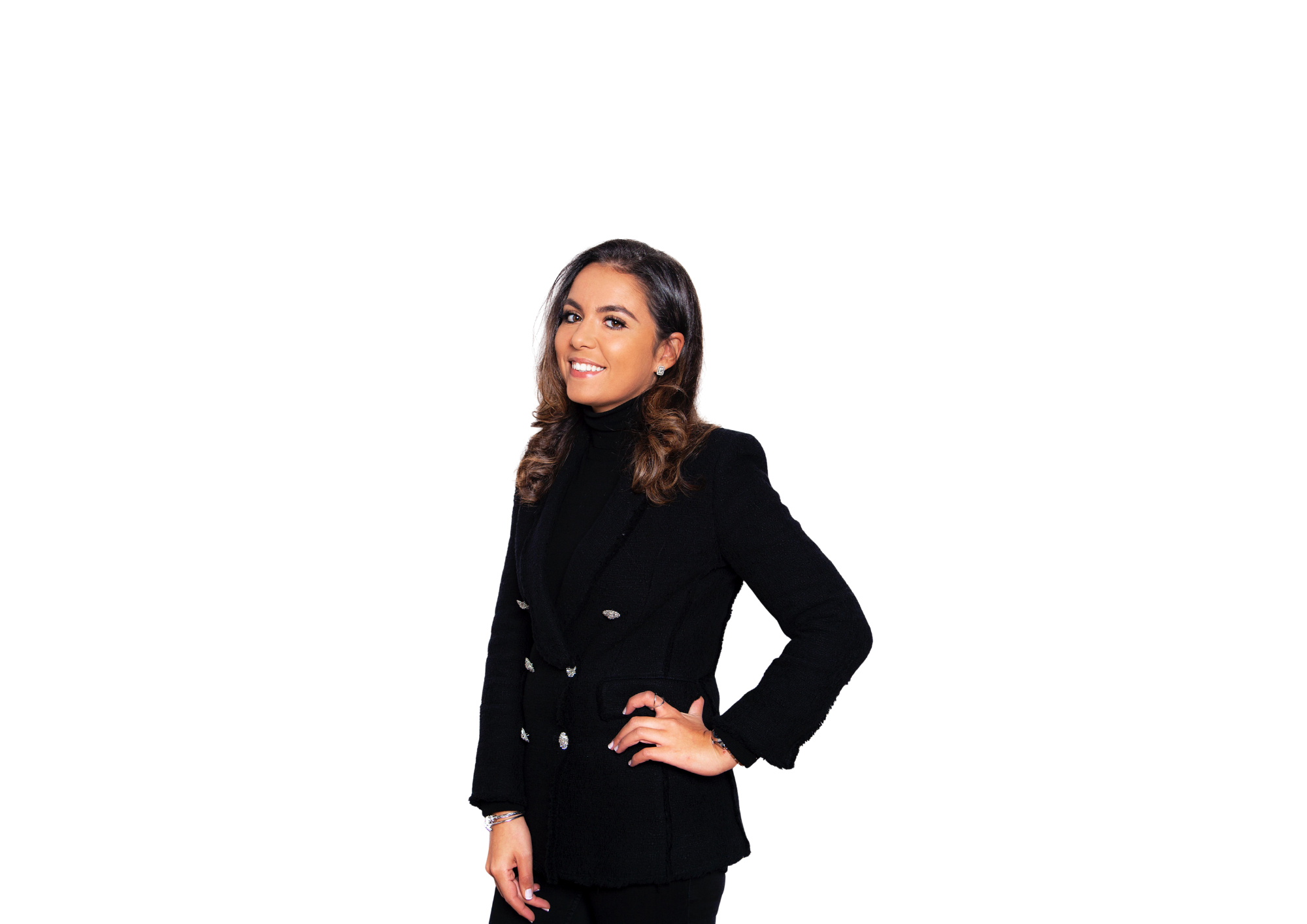 Une femme souriante avec des cheveux bruns ondulés, portant un costume noir avec des boutons argentés, pose avec une main sur la hanche