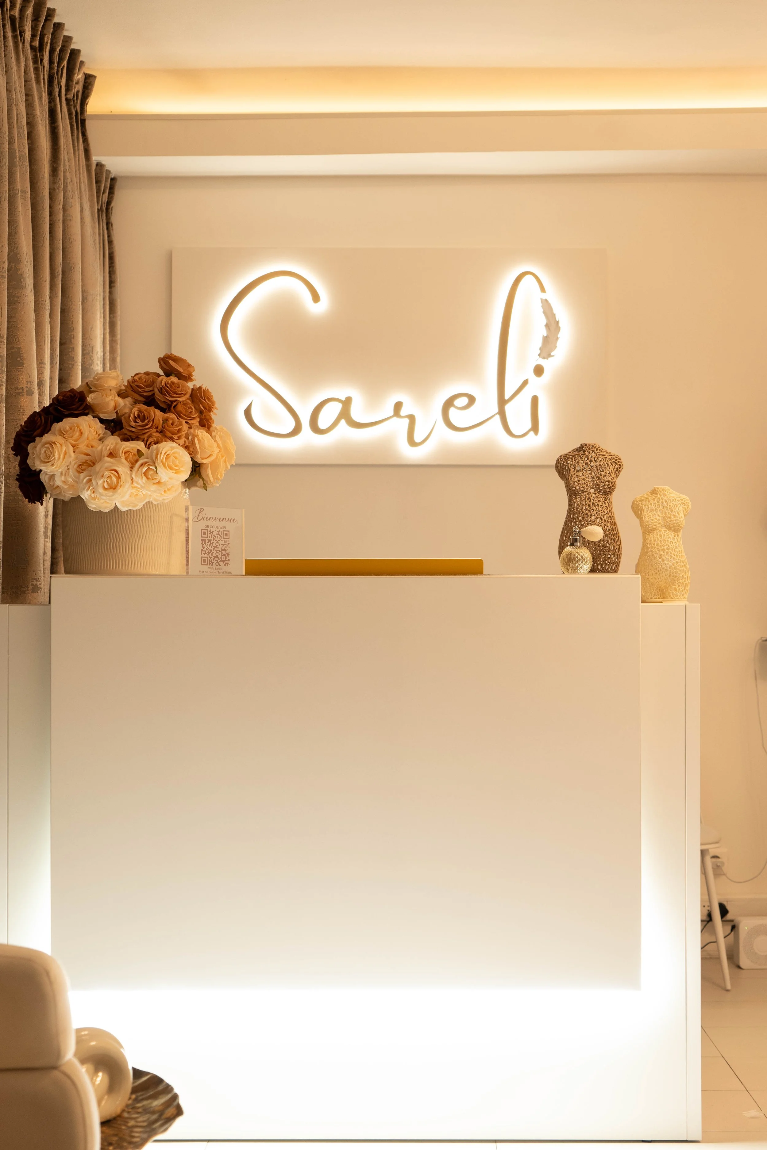 Panneau lumineux avec le nom 'Sareli' en script, bouquet de roses dans un vase blanc, deux bustes en relief en léopard, figurine en verre, et décorations en forme de bustes sur un comptoir blanc dans une réception.